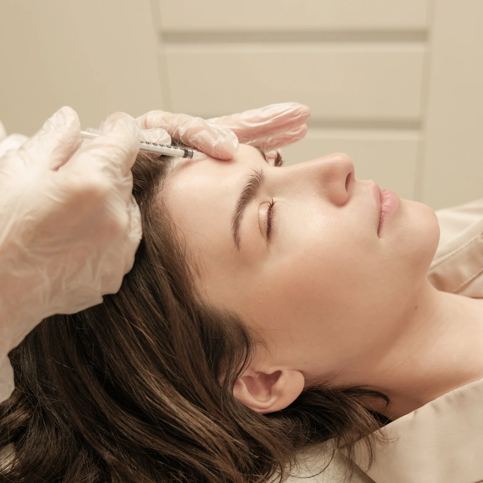 A woman receiving a cosmetic injection in her forehead while lying down with her eyes closed at a medical clinic.
