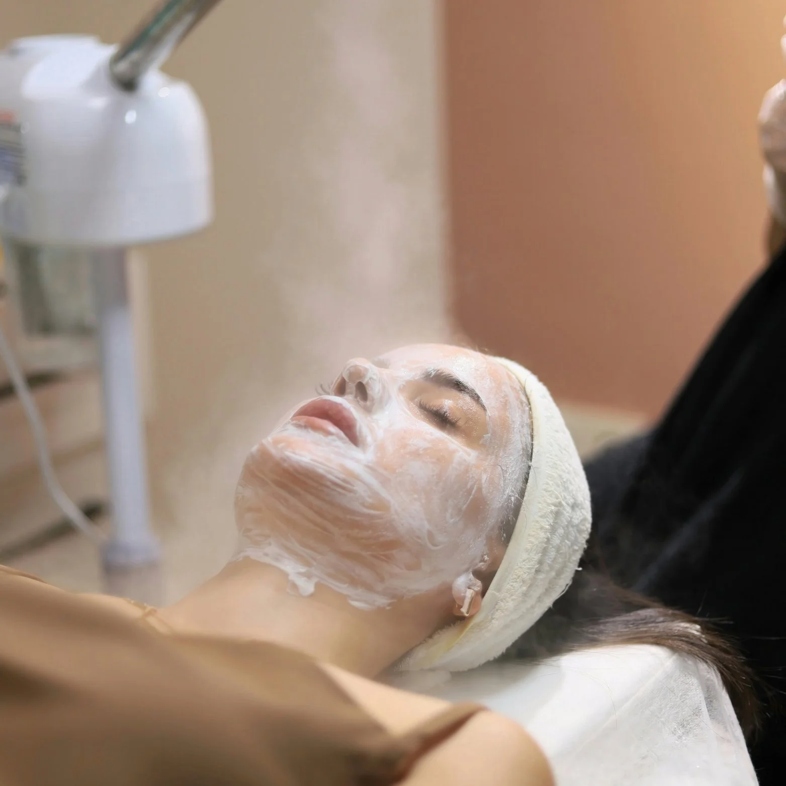 A woman lying on a spa bed with her eyes closed, receiving a facial treatment with a white facial mask, in a skincare clinic or spa setting.