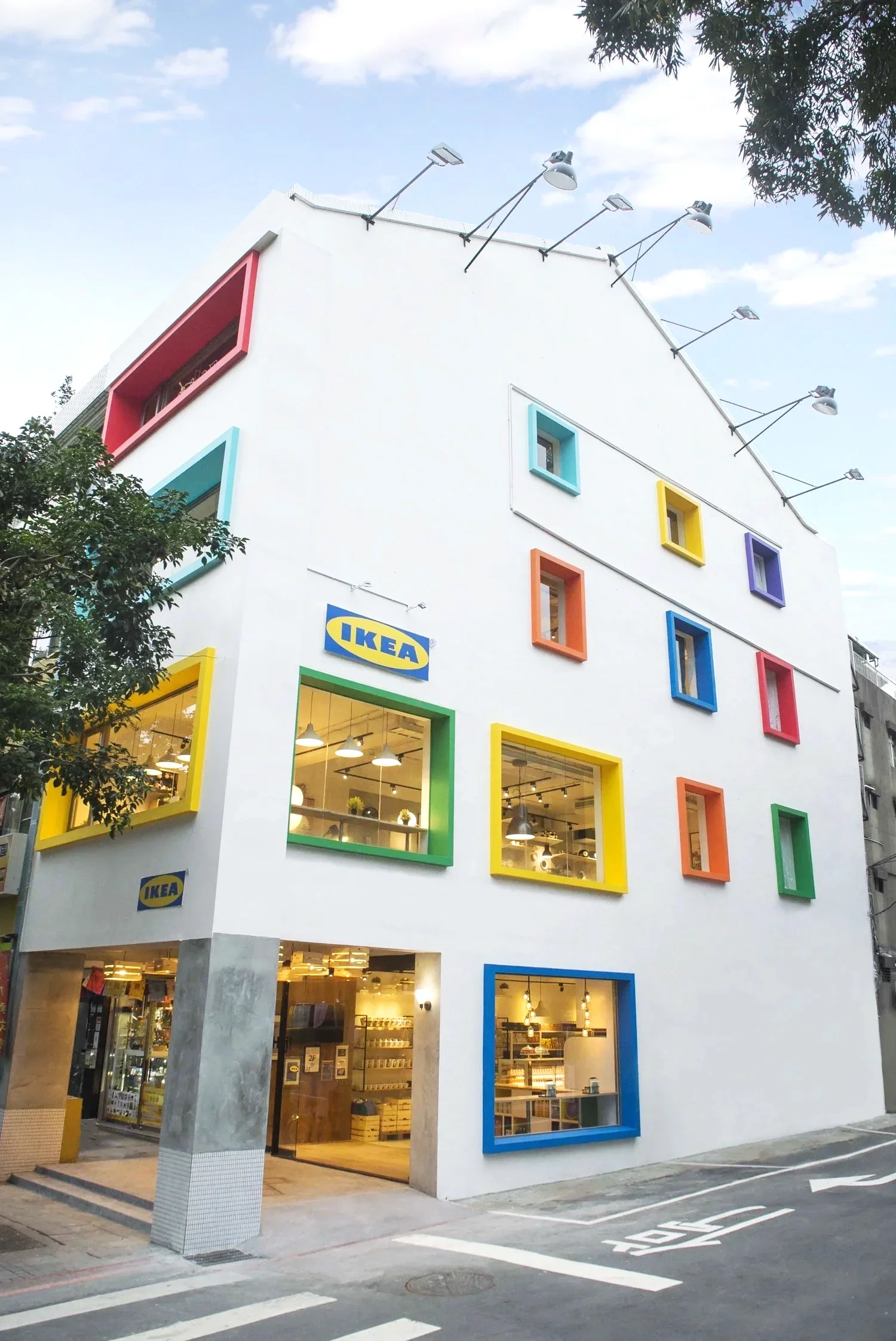 Exterior of a white building with colorful window frames in blue, red, yellow, purple, green, orange, and teal, housing an IKEA store. The store's sign is visible near the entrance, and the interior lights can be seen through large windows. The building is on a street with a crosswalk and parking space.