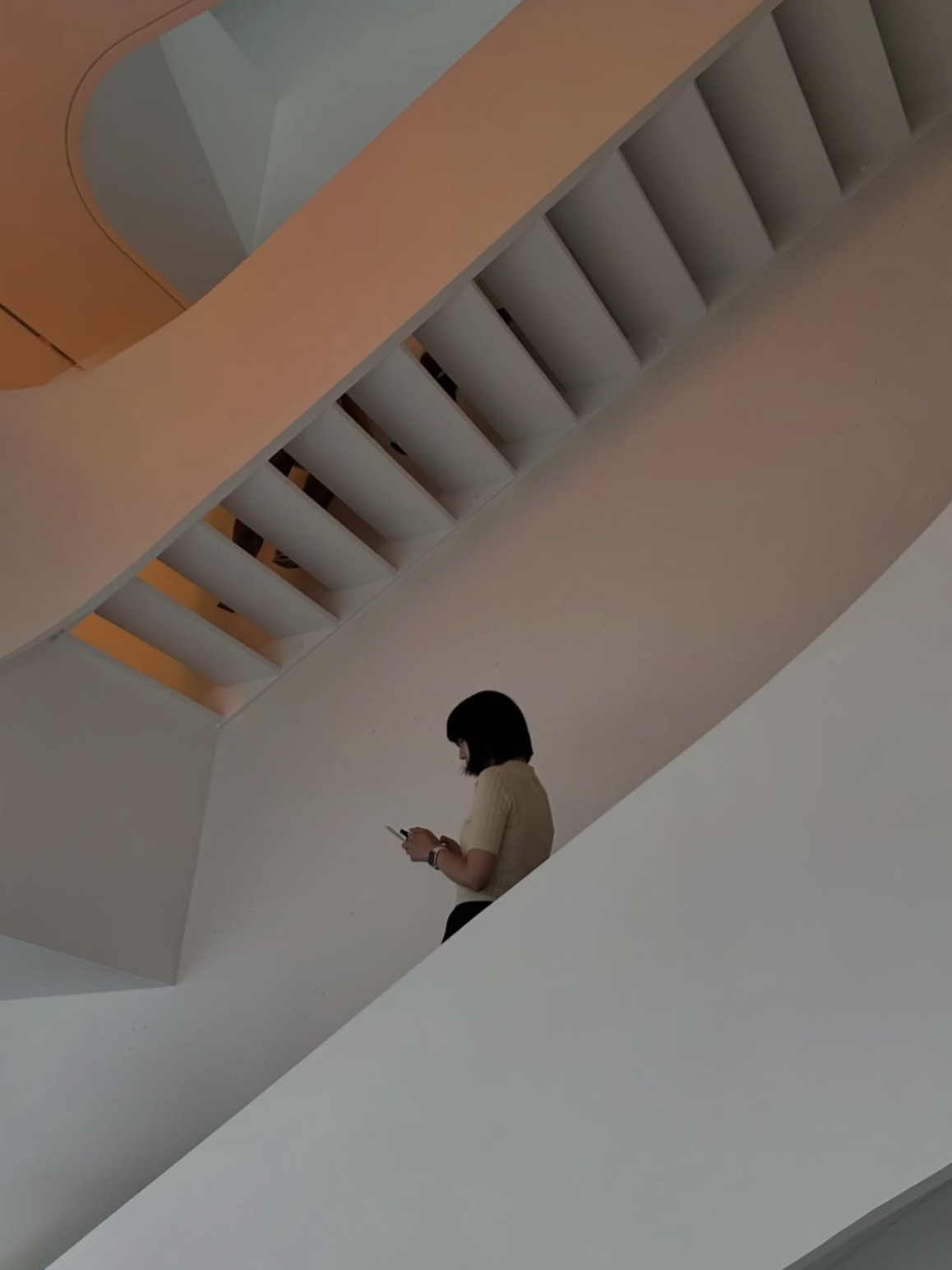 Une femme avec une chemise beige qui regarde son téléphone tout en étant assise sur une structure blanche dans un espace intérieur moderne avec des escaliers en arrière-plan.