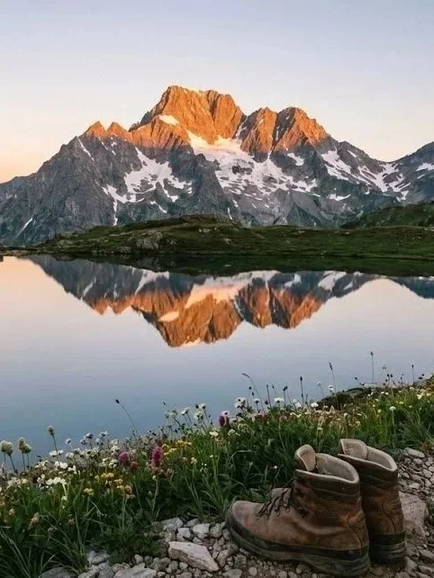 Montagnes enneigées au coucher du soleil, reflet dans un lac calme, chaussures de randonnée sur le sol rocailleux avec des fleurs en premier plan.