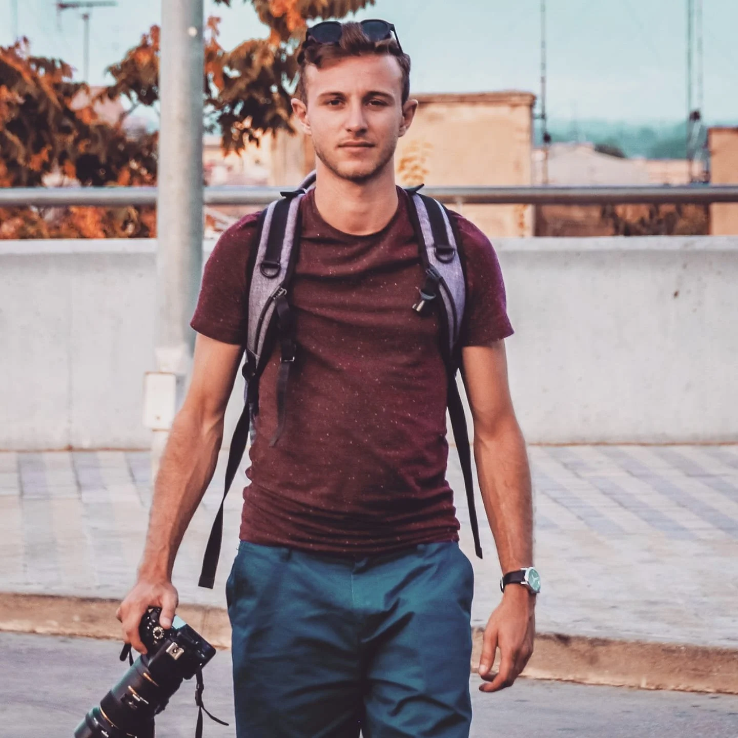 Jeune homme avec un sac à dos et une caméra, debout à l'extérieur, avec des arbres aux feuillesorange et une barrière en arrière-plan.