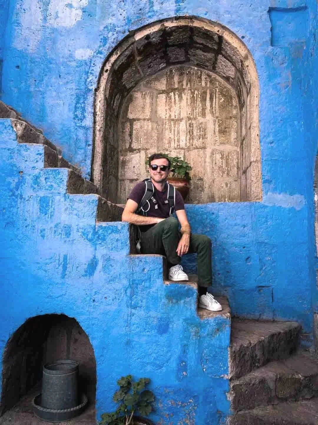 Homme assis sur des escaliers bleus, portant un sac à dos, lunettes de soleil, avec un mur en pierre et un pot de fleurs en arrière-plan.
