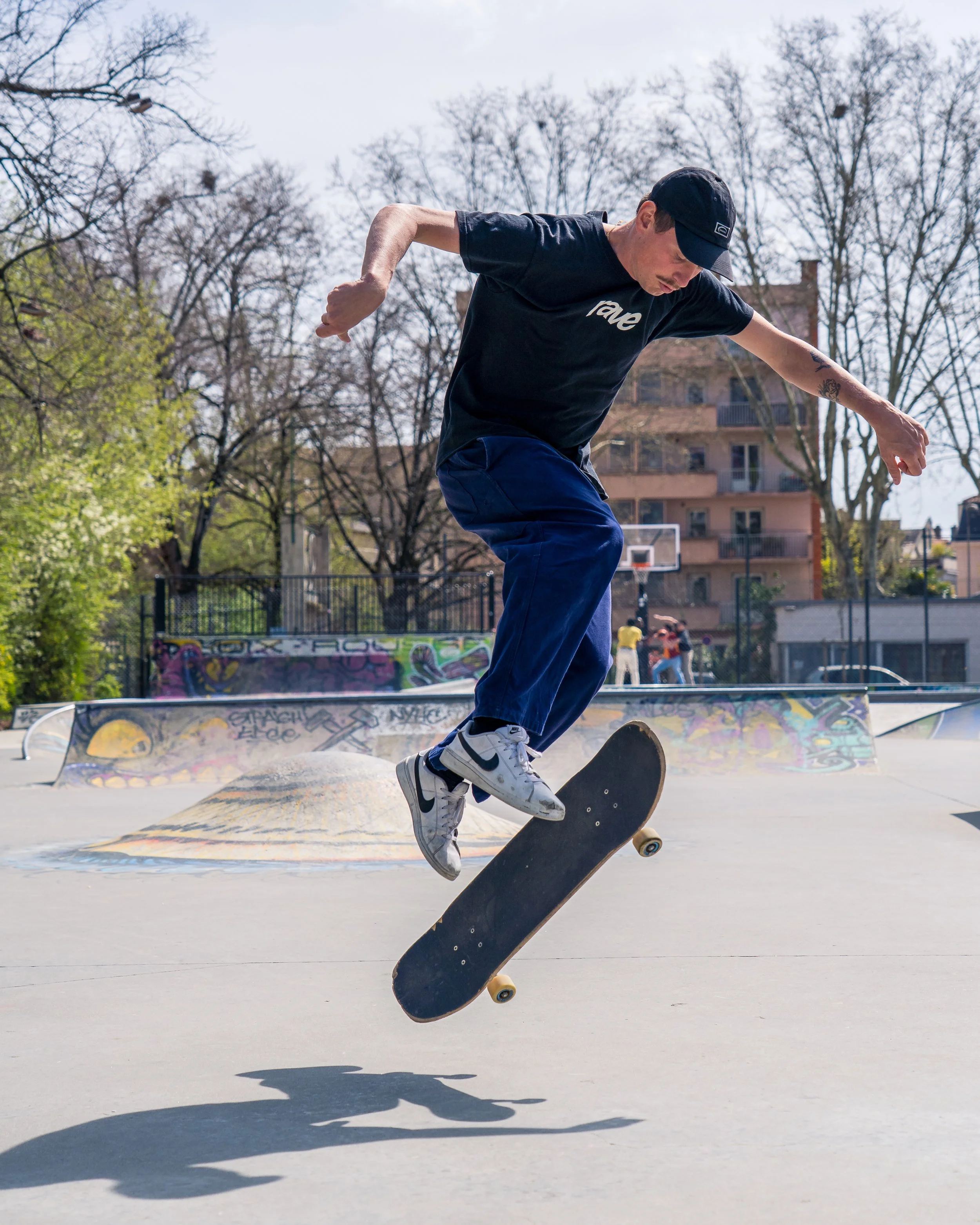 Un jeune homme en t-shirt noir, pantalon bleu et casquette noire fait du skateboard dans un skatepark en plein air par une journée ensoleillée avec des arbres en arrière-plan.