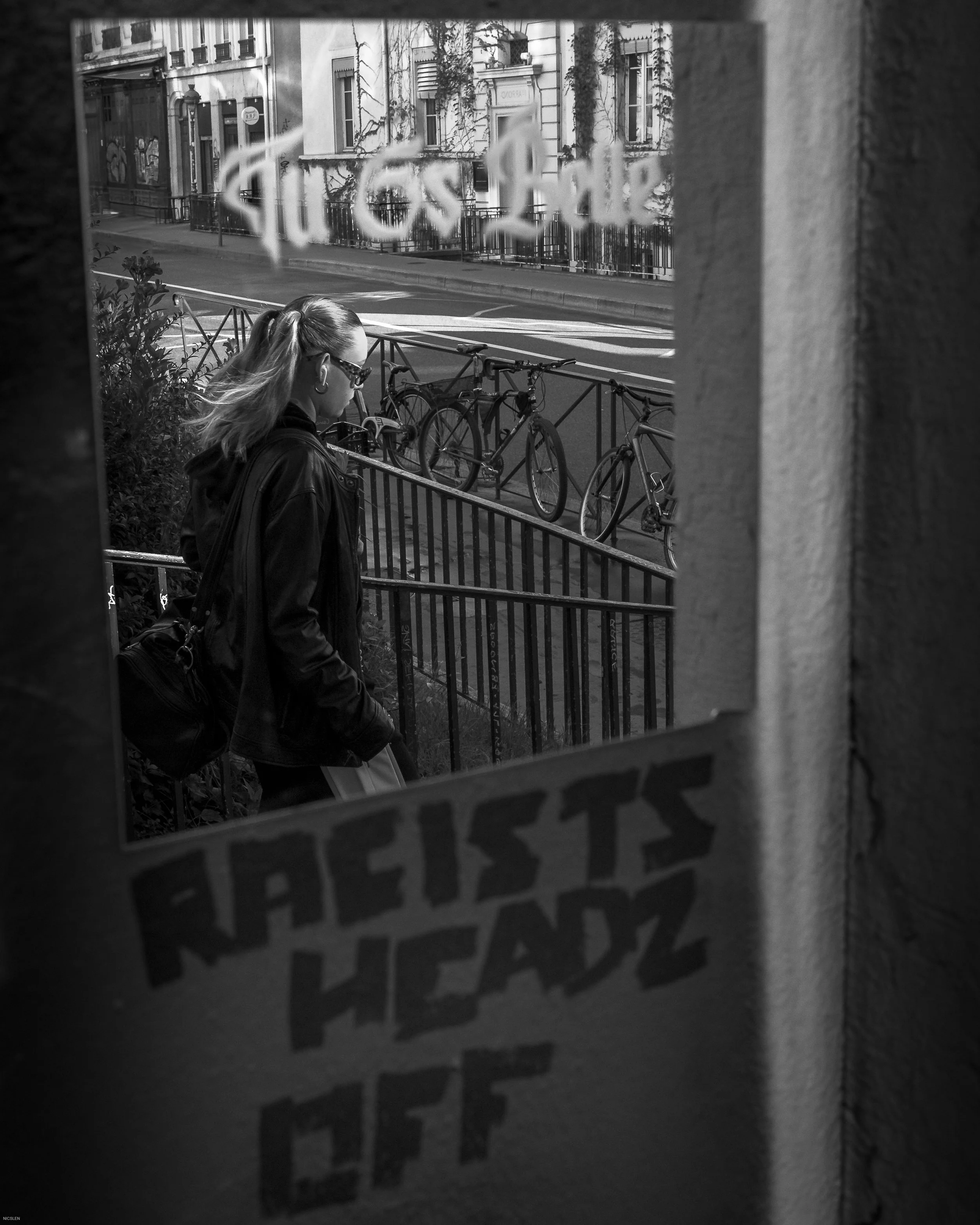 Une femme marche dans une rue parisienne, vue par une fenêtre avec des graffitis, entourée de vélos stationnés et de bâtiments en arrière-plan.
