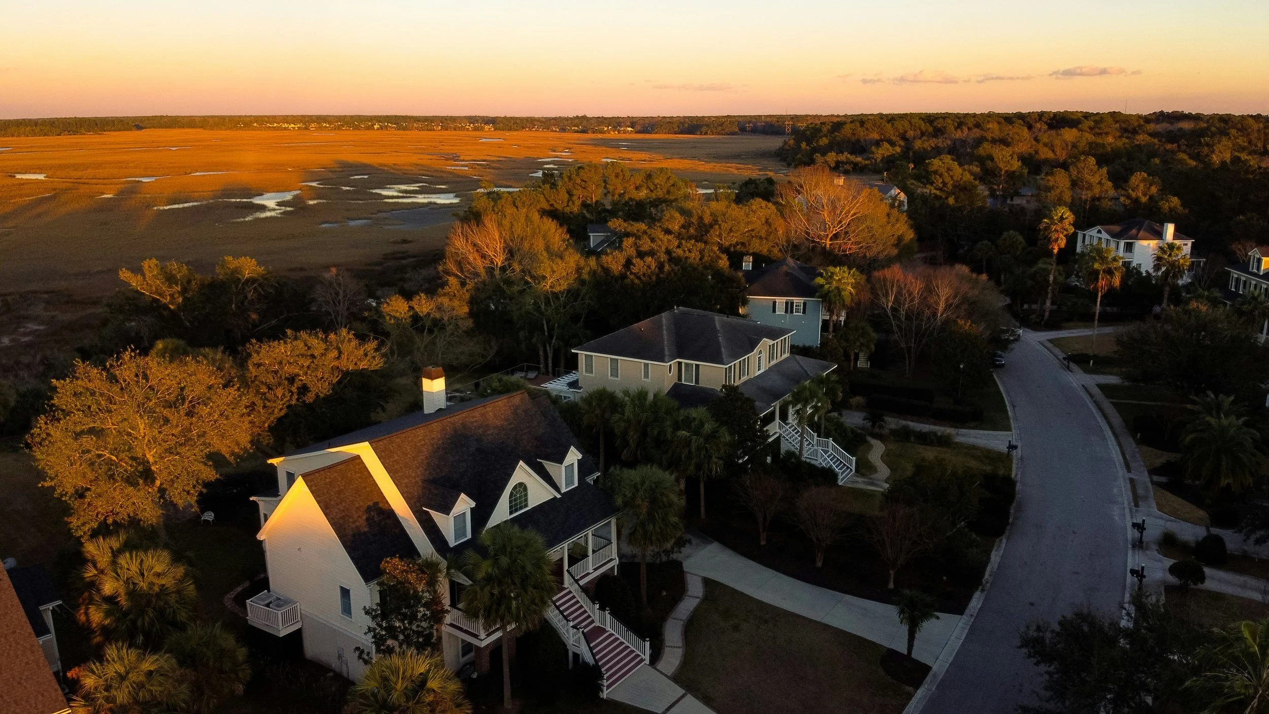 An aerial view of a neighborhood at sunset, showing houses surrounded by trees and palm trees, with a large expanse of marshland or wetland in the distance under a pink and orange sky.