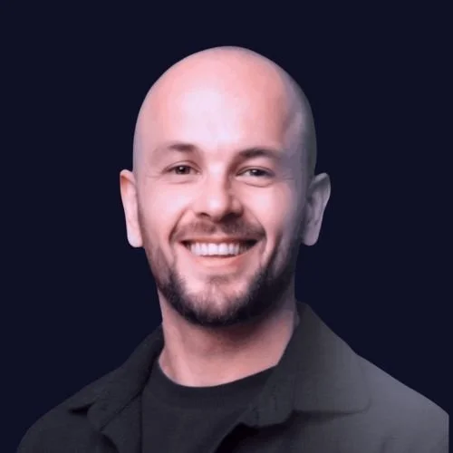 A smiling man with a bald head and beard, wearing a black jacket against a dark background. Our Founder Alex Elnaugh.
