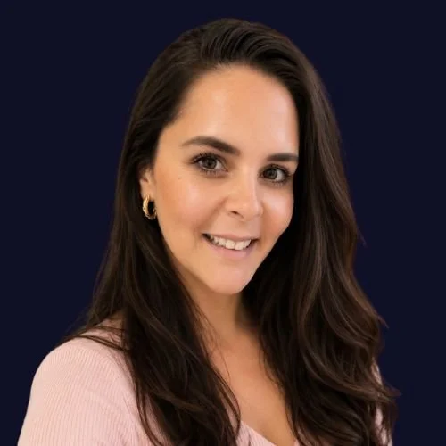 A young woman with long dark hair, wearing a light pink top and gold earrings, smiling at the camera against a dark background. Our Co Founder Emma Pringle.