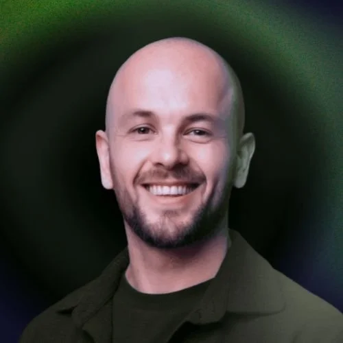 A smiling bald man with a beard, wearing a dark jacket, against a blurred green and dark background.