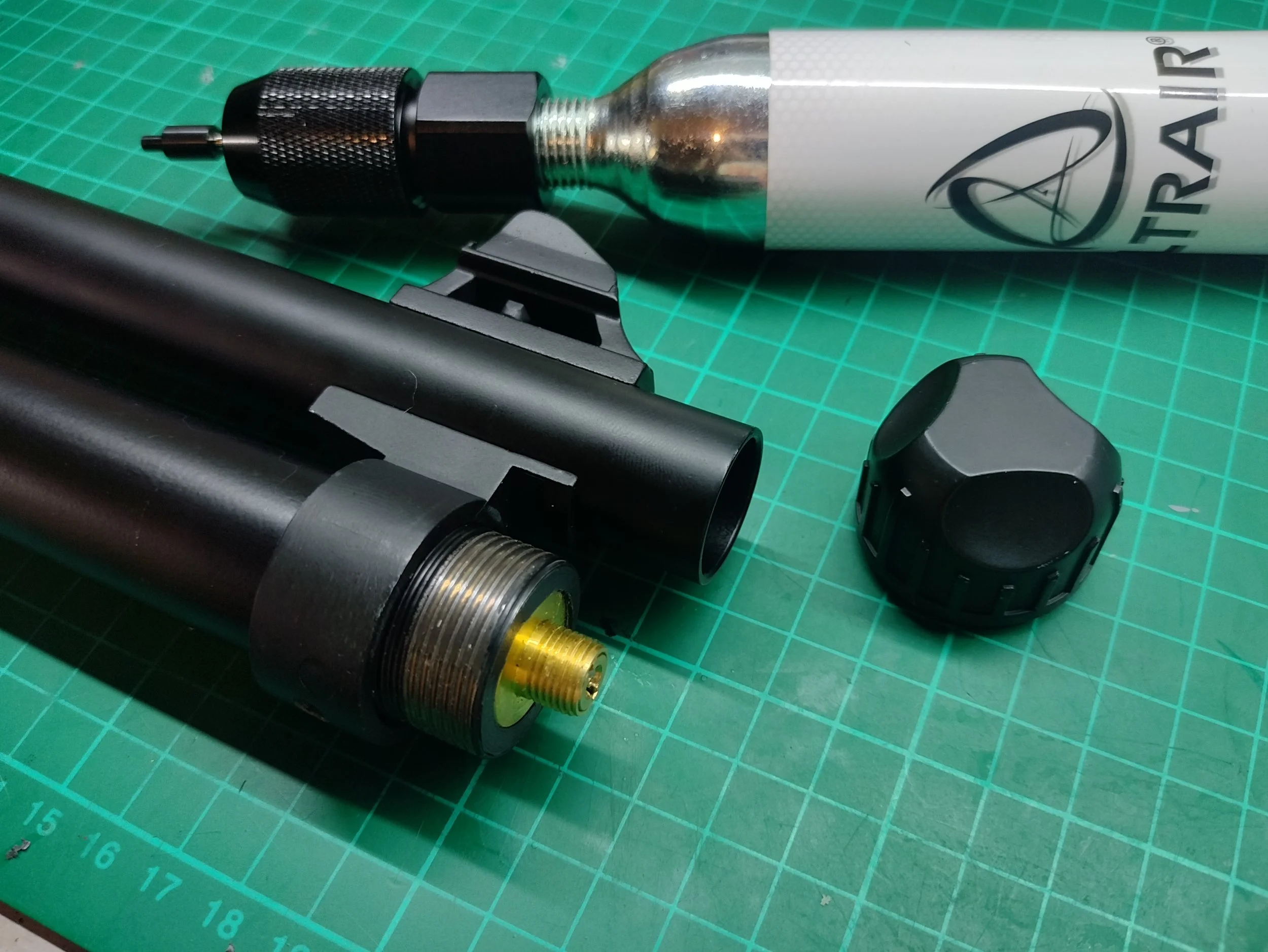 A airsoft tri-shot shotgun with a CO2 charging nozzle, and a canister of compressed CO2 on a green cutting mat, along with a black end cap.