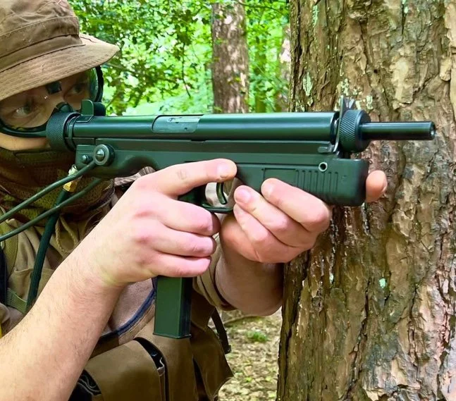 Person wearing camouflage gear and glasses aiming a black airsoft submachine gun at a tree in a forest.