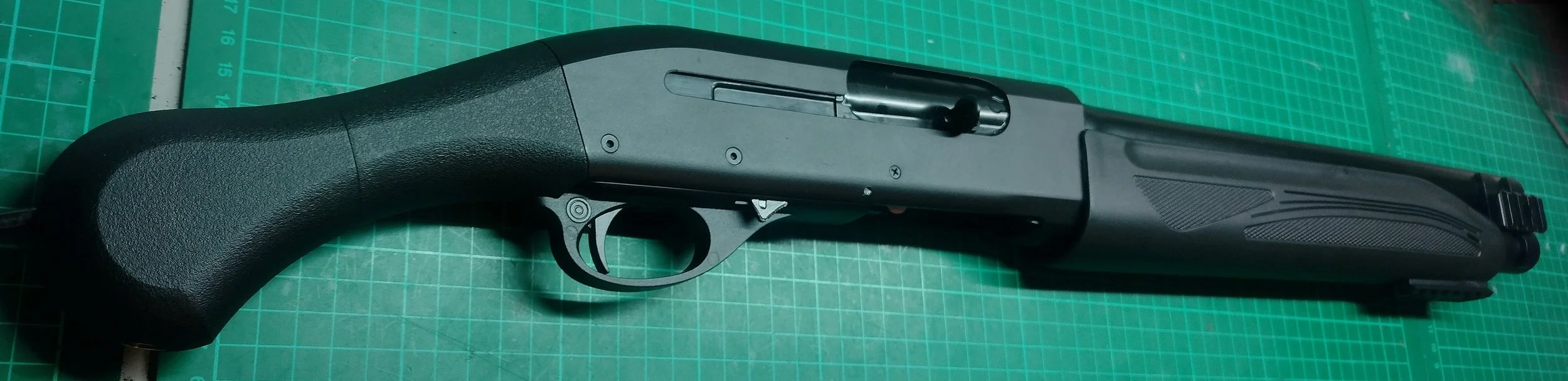 Black airsoft HPA shotgun placed on a green cutting mat with measurement grid.