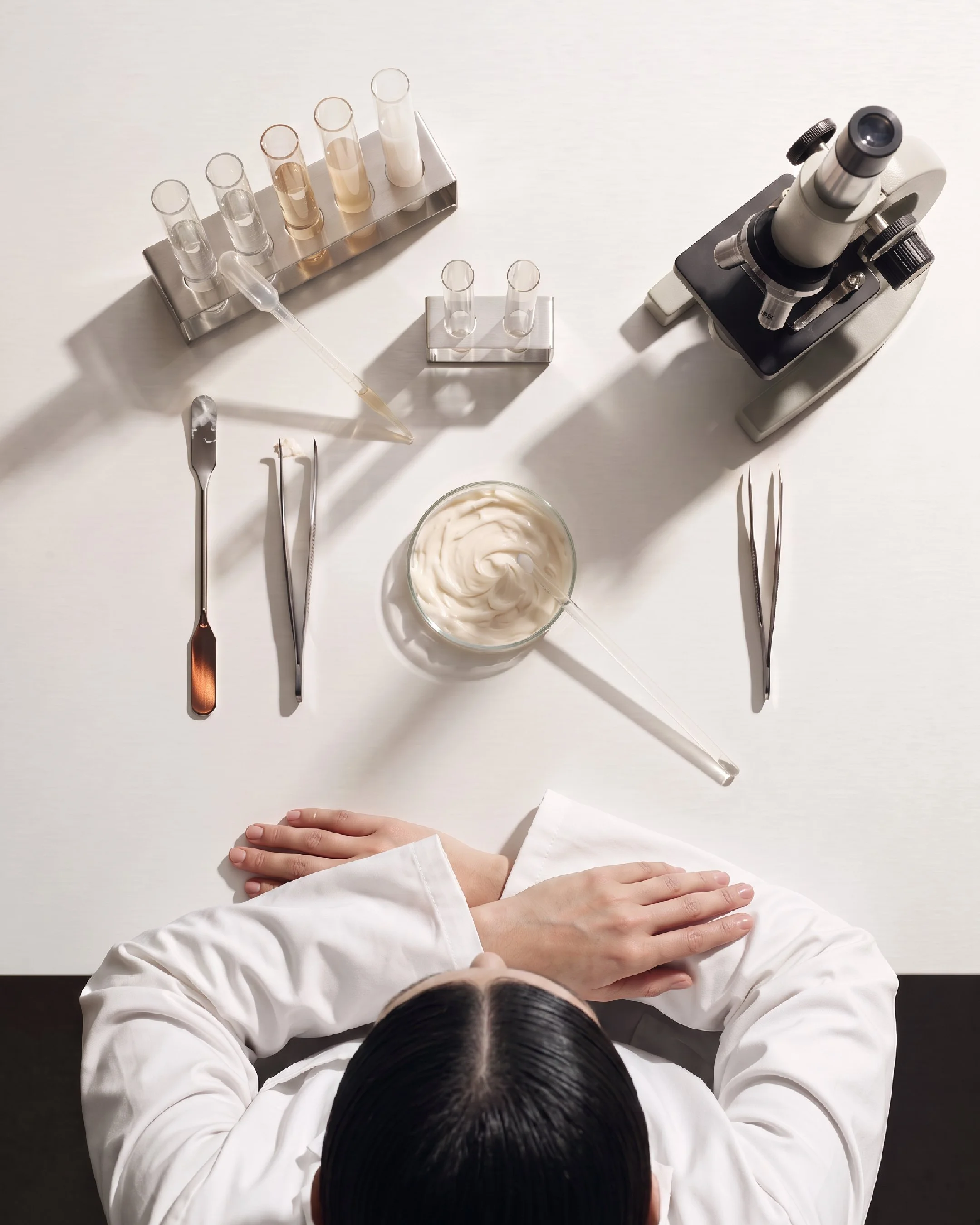 Ai generet laboratorie univers. Kampagne materiale skabt af projekt 01. En person iført en hvid laboratoriefrakke, der sidder med armene krydset foran på et blankt hvidt bord, omgivet af laboratorieudstyr såsom reagensglas, pipetter og en mikroskop.