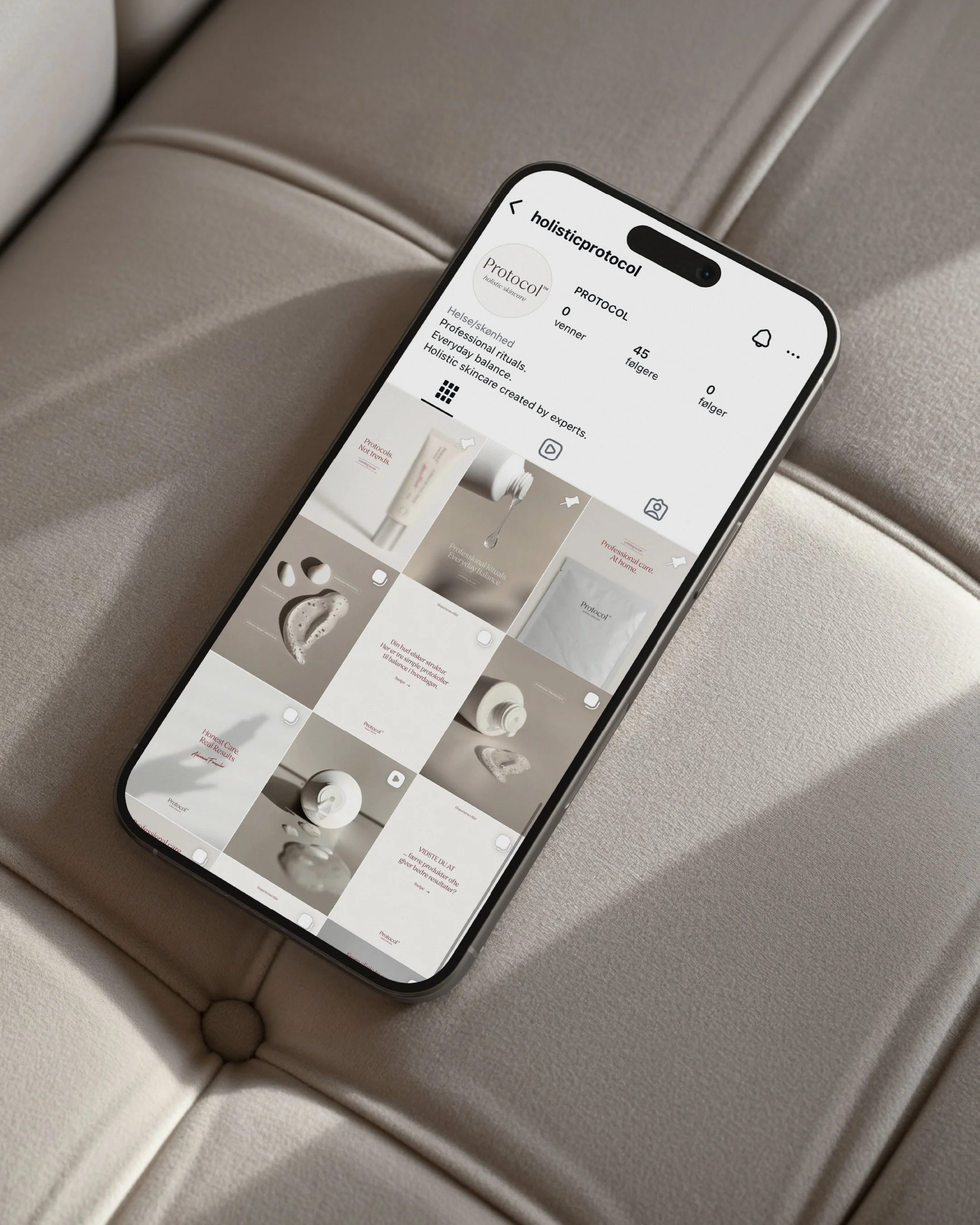 En smartphone på en beige puf, der viser en app kaldet 'holisticprotocol' med et profilbillede og punkter af opslag, produkter og information på skærmen.