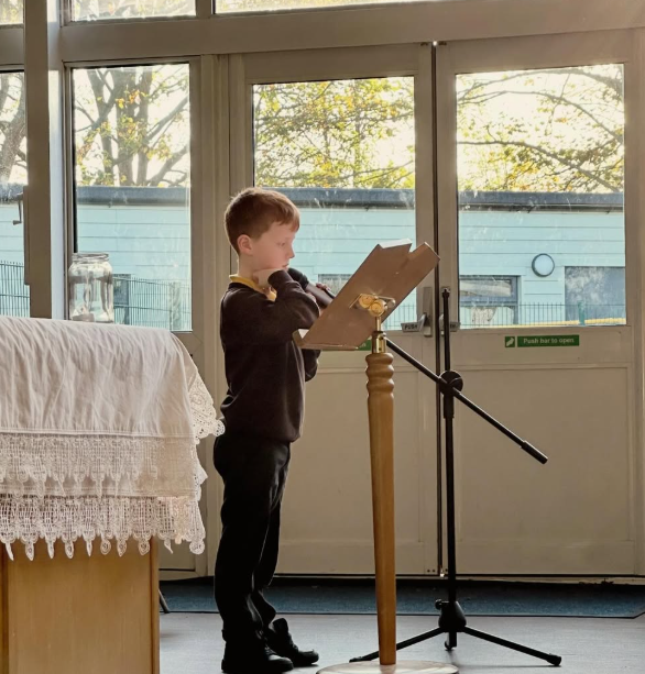 A young boy in school uniform reading aloud from a book on a wooden stand, using a microphone in a room with large windows and a door decorated with a lace tablecloth on a nearby piece of furniture.