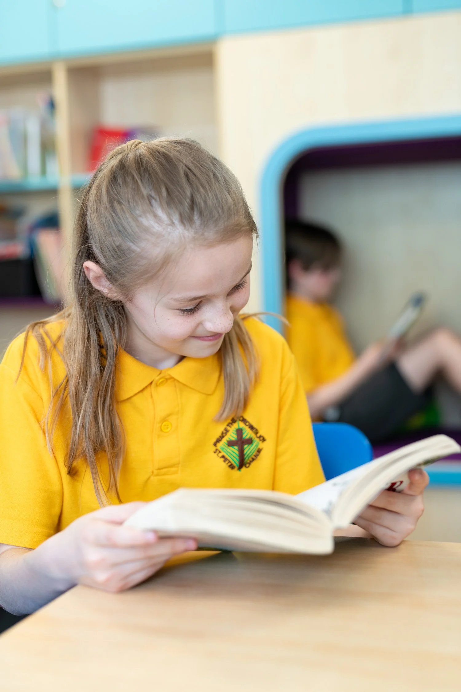 A young girl with long, light brown hair wearing a yellow shirt reading a book in a classroom.