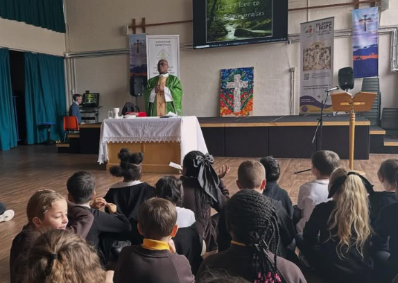 A woman speaks to a group of children seated on the floor in a church or school hall, with a cross and religious banners in the background.