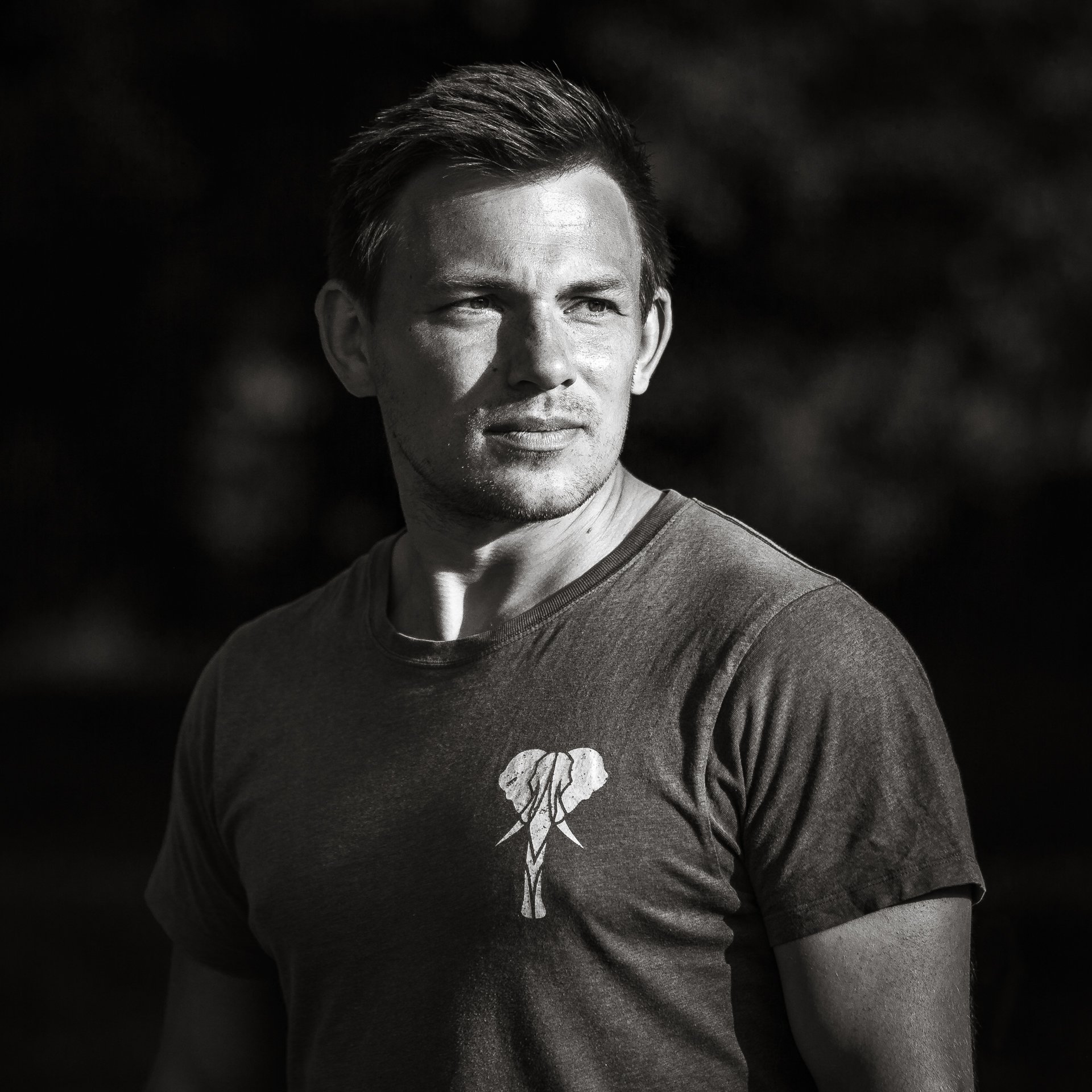 A young man with short hair looking to the right outdoors, wearing a t-shirt with an elephant graphic.