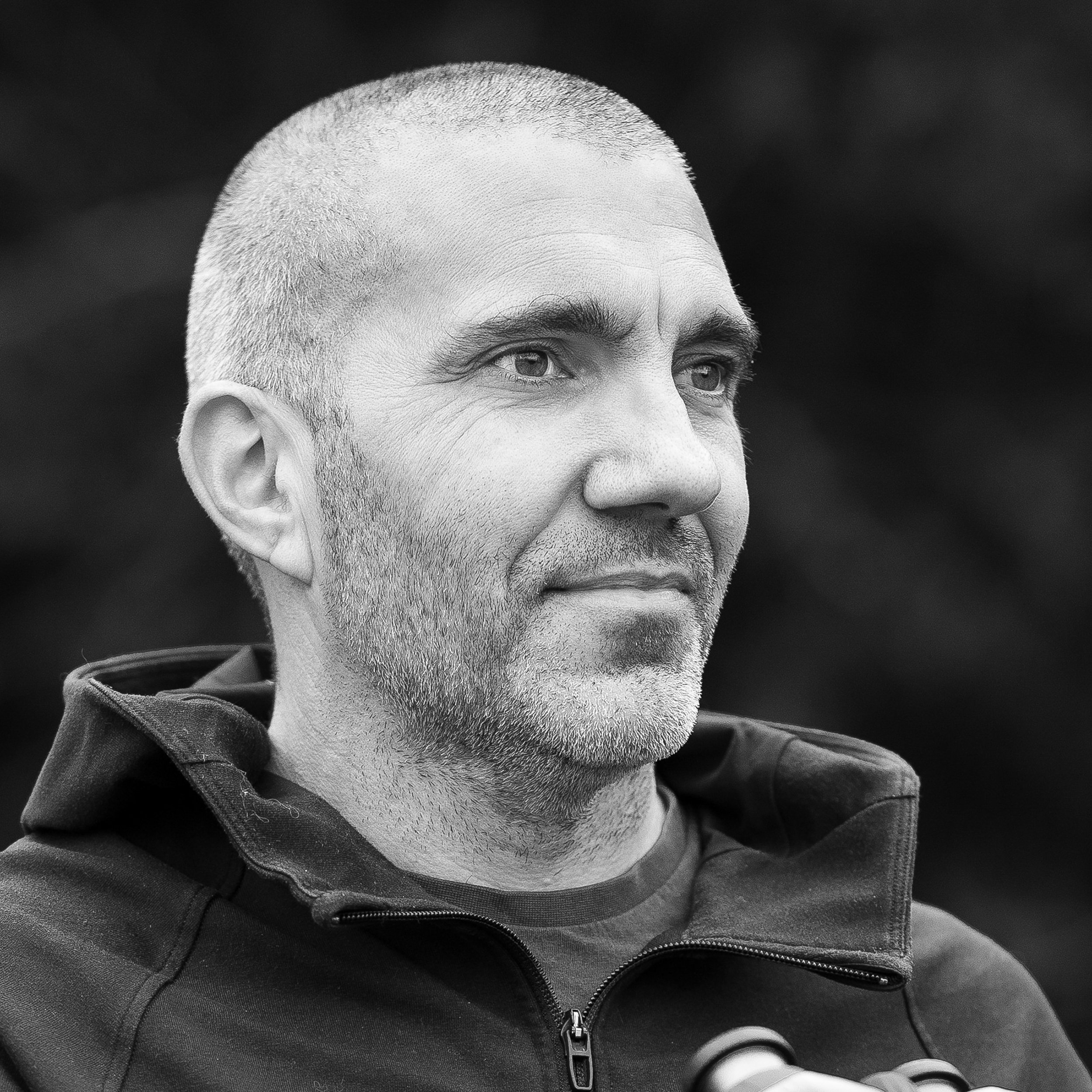 Black and white photo of a middle-aged man with short, light hair and stubble, wearing a zip-up jacket, looking to the side with a calm expression.