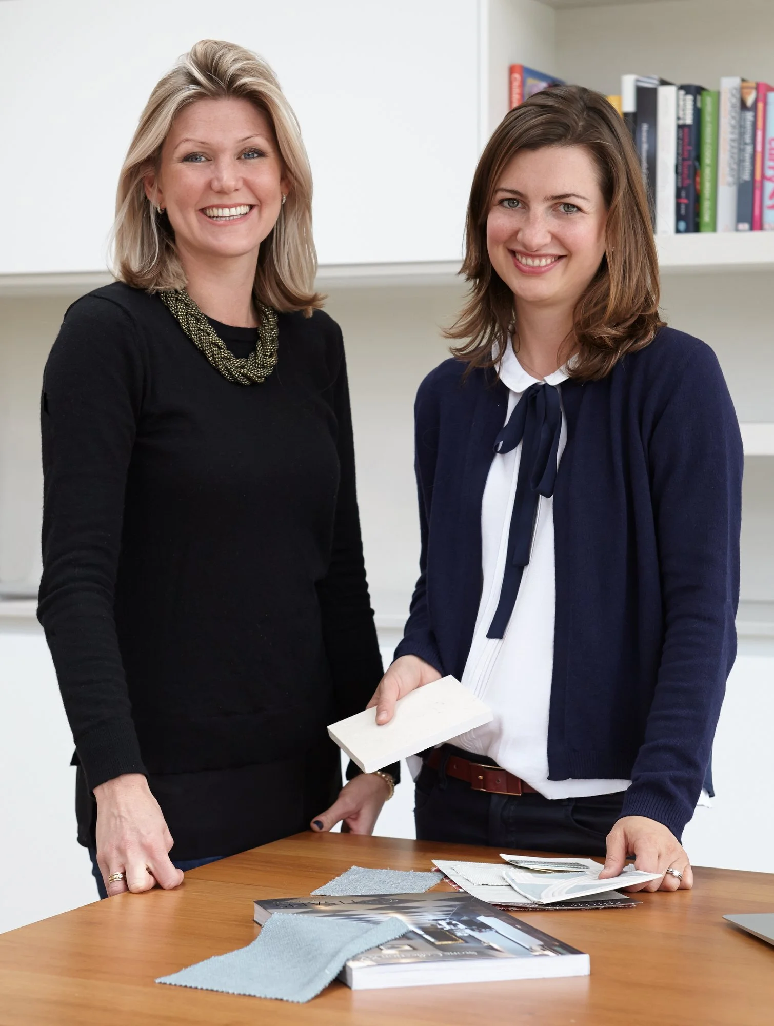 Two interior designers from Schiller Beynon Interior Design  standing at a table, holding a sample of material, with books and fabric swatches on the table, in an office or design studio setting.