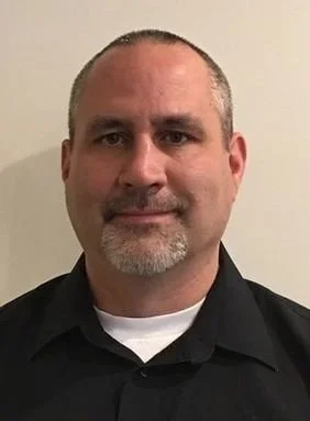 Close-up of a man with short hair and a beard, wearing a black collared shirt, against a plain background.