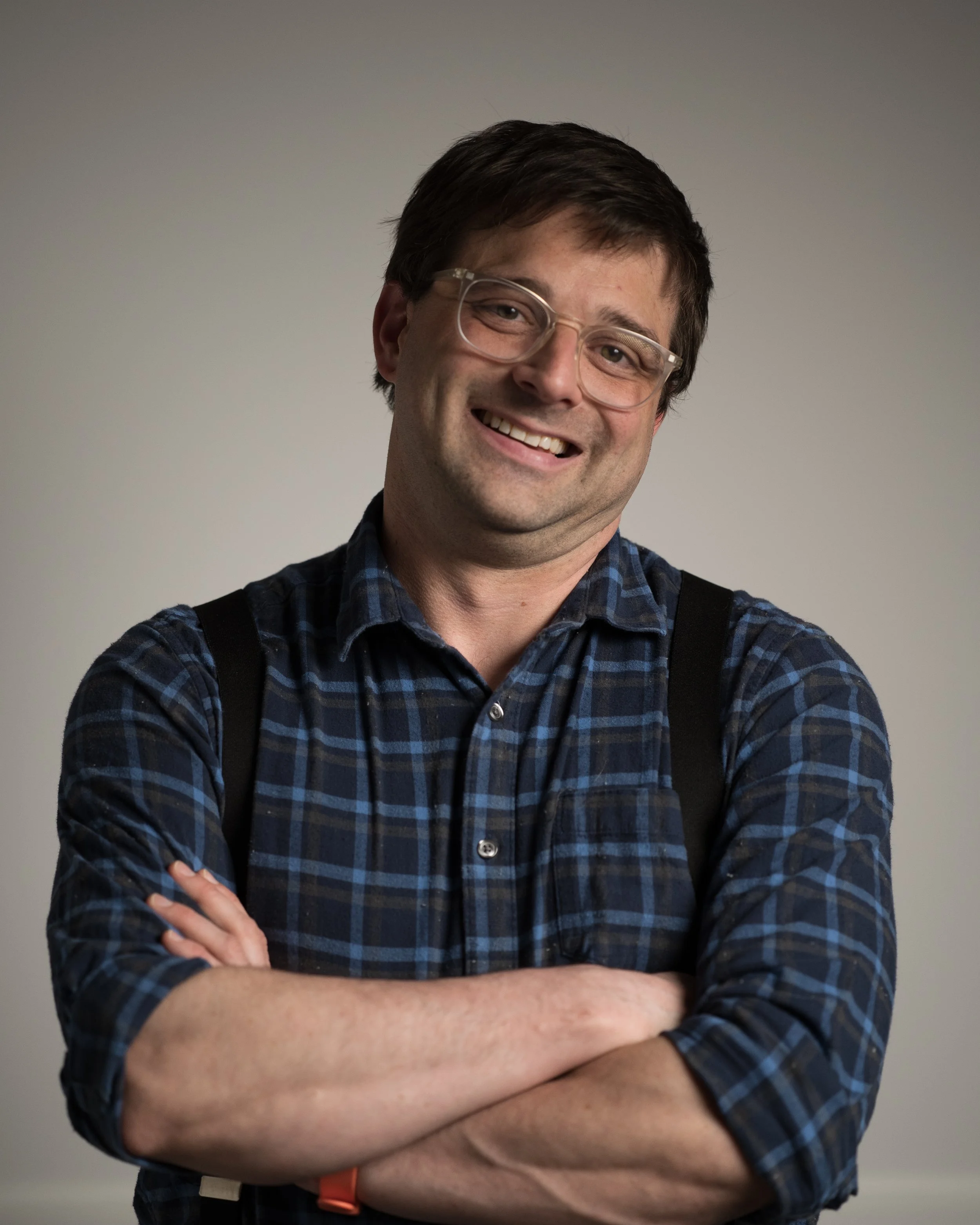 A smiling man with glasses wearing a dark blue plaid shirt with rolled-up sleeves and a backpack, crossing his arms against a plain background.