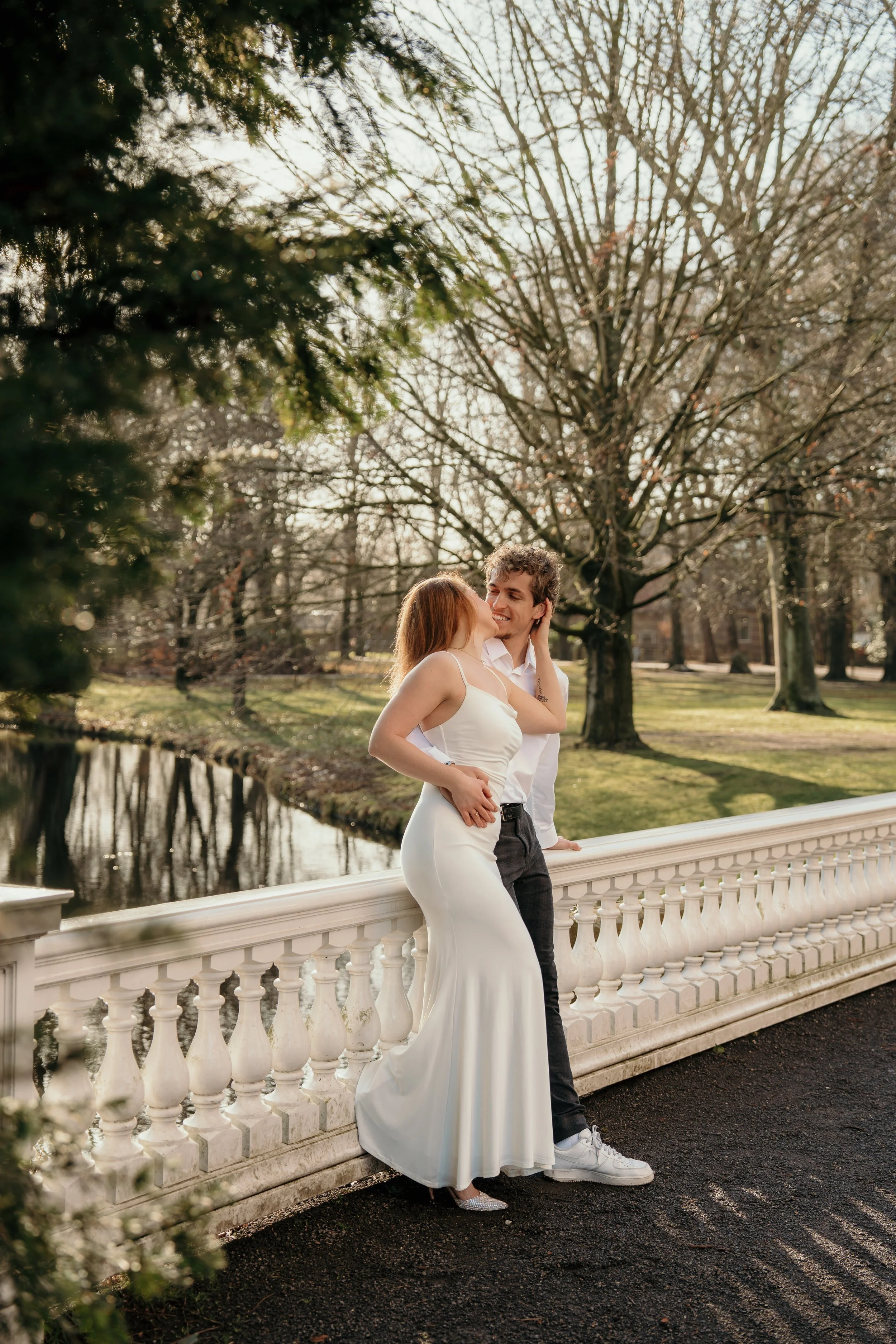 Een bruidspaar dat op een witte balustrade staat in een park met bladeren en bomen, de bruid draagt een witte trouwjurk en de man een witte overhemd en donkere broek, ze lachen naar elkaar terwijl de zon ondergaat tijdens een bruidsreportage van Rosa
