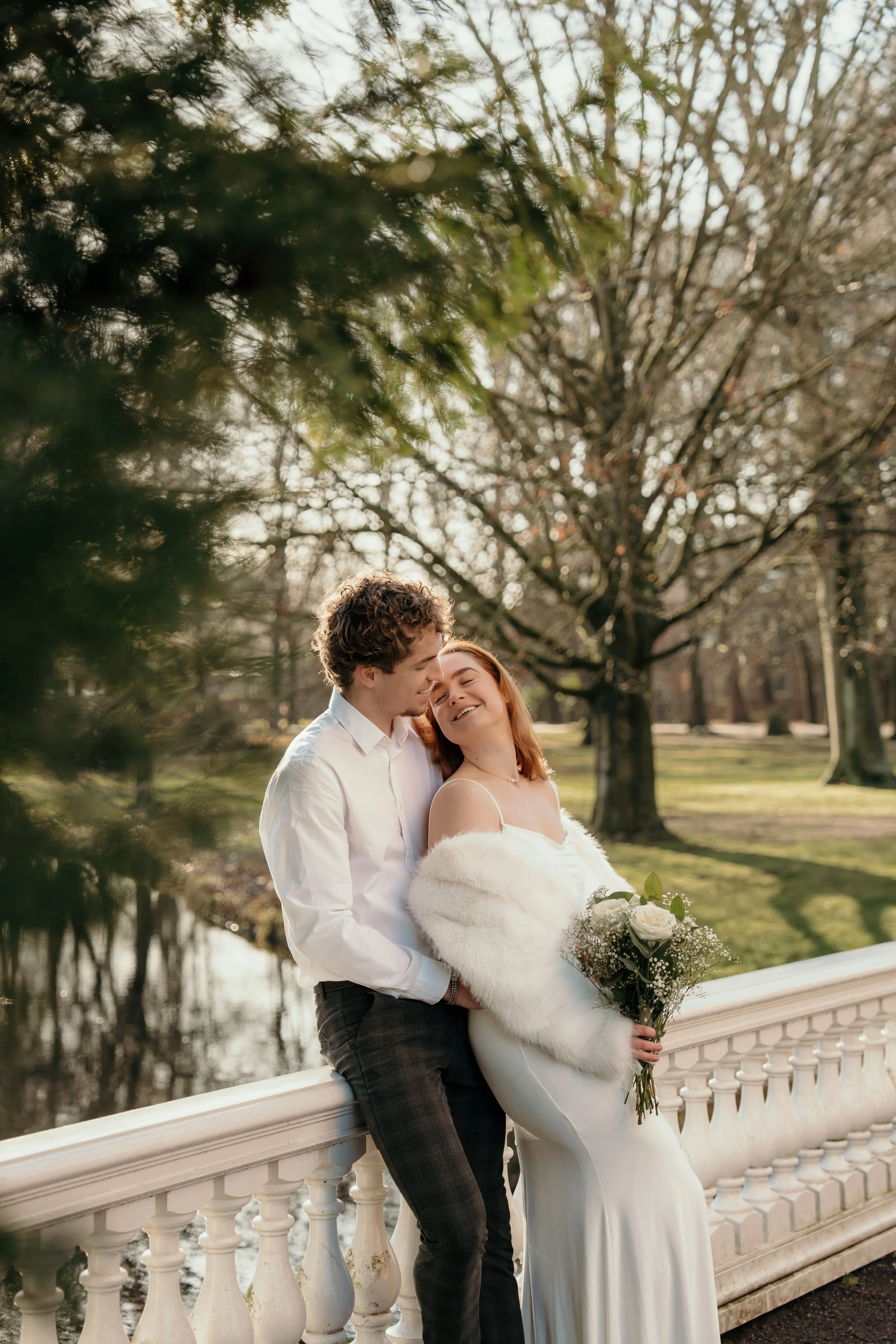 Een bruidspaar op hun bruiloft zittend op een witte balustrade bij een park, omringd door bomen en een rustig water. Ze glimlachen en lijken gelukkig tijdens een bruidsreportage van Rosa Lynn Signer.