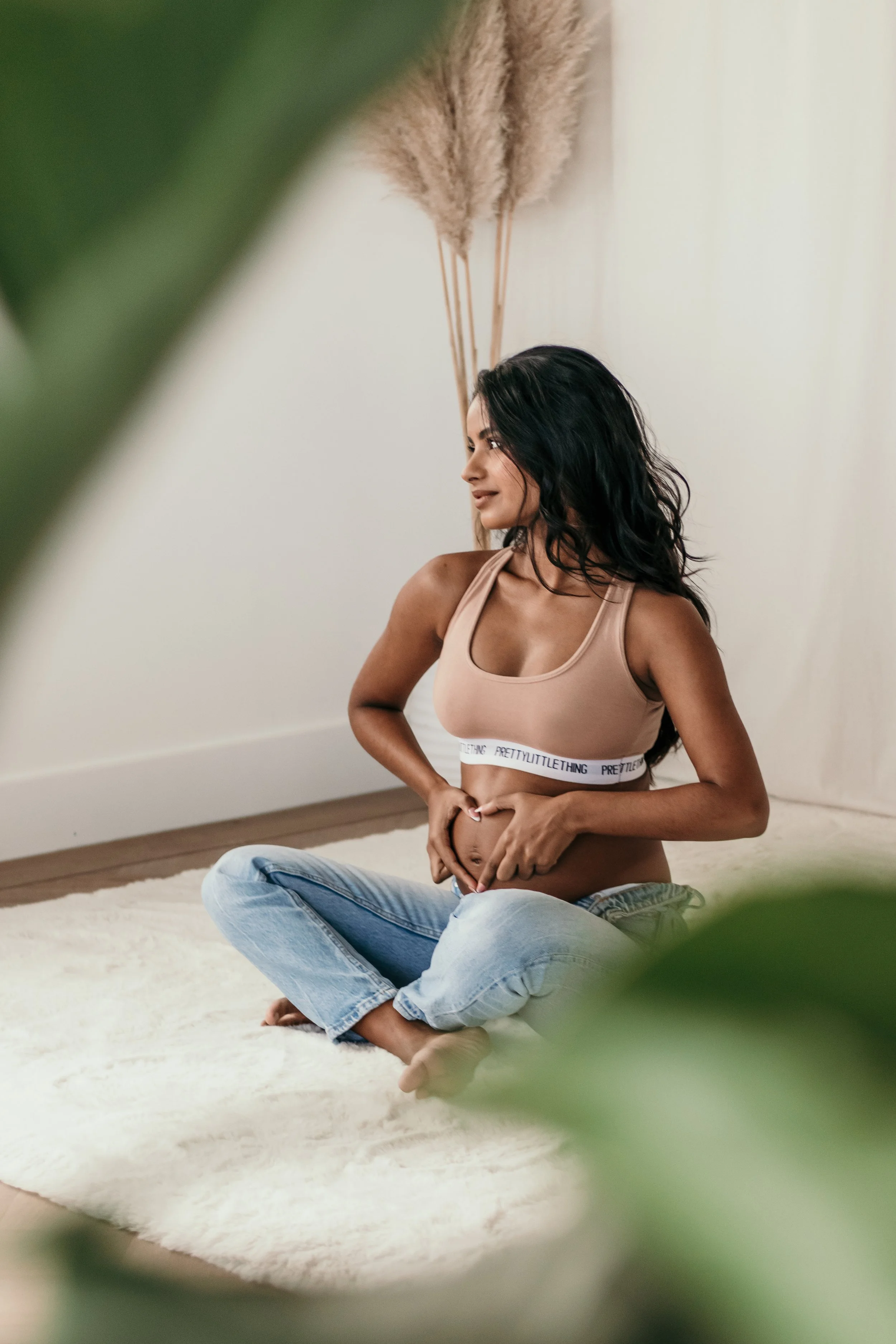 Een zwangere vrouw zit op een witte tapijt, met haar handen op haar buik, terwijl ze glimlacht tijdens een zwangerschapsshoot van Rosa Lynn Signer in een daglichstudio.