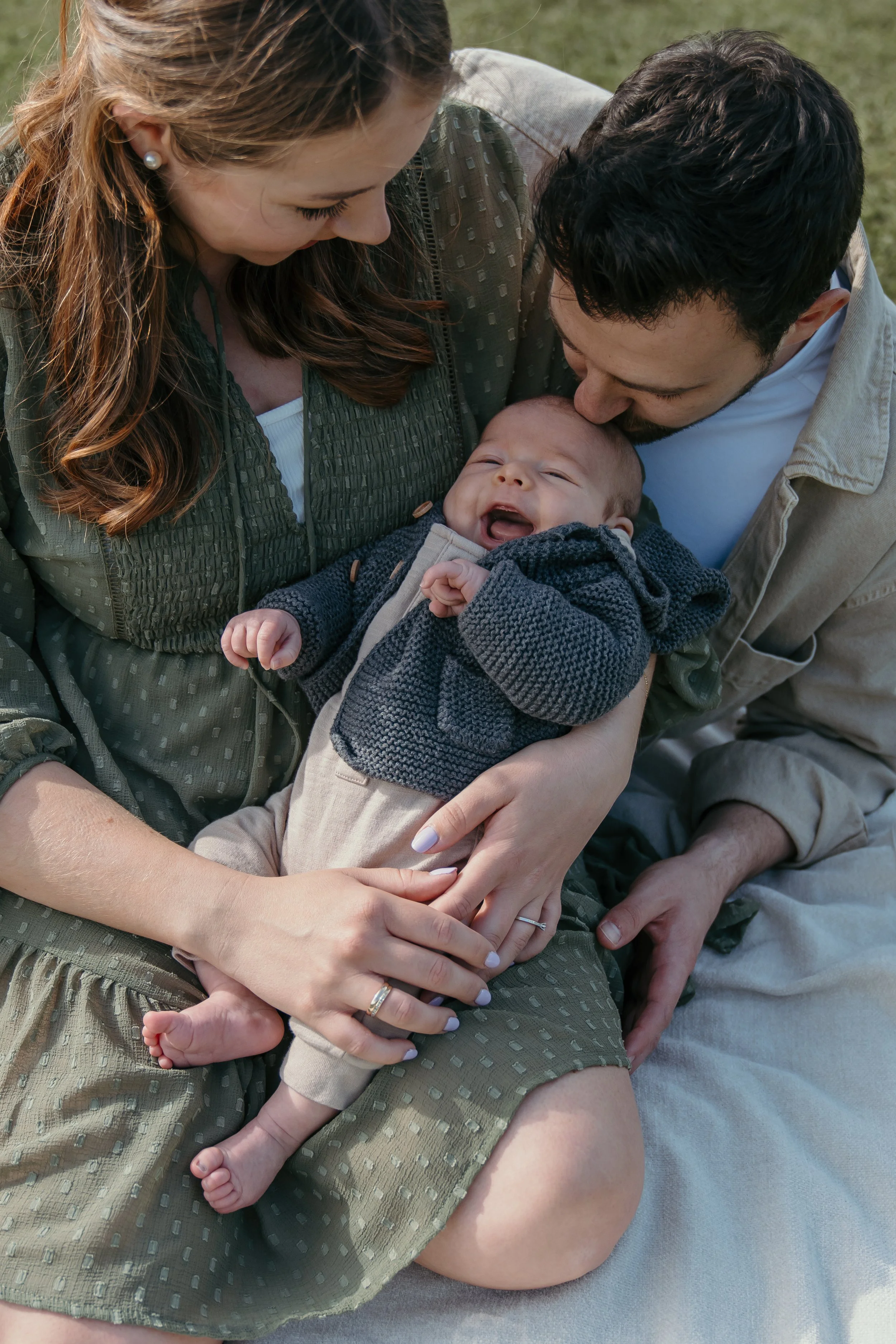 Een jong gezin met een baby zit samen buiten op het gras, ze lachen en knuffelen de baby tijdens een new born shoot van Rosa Lynn Signer. 