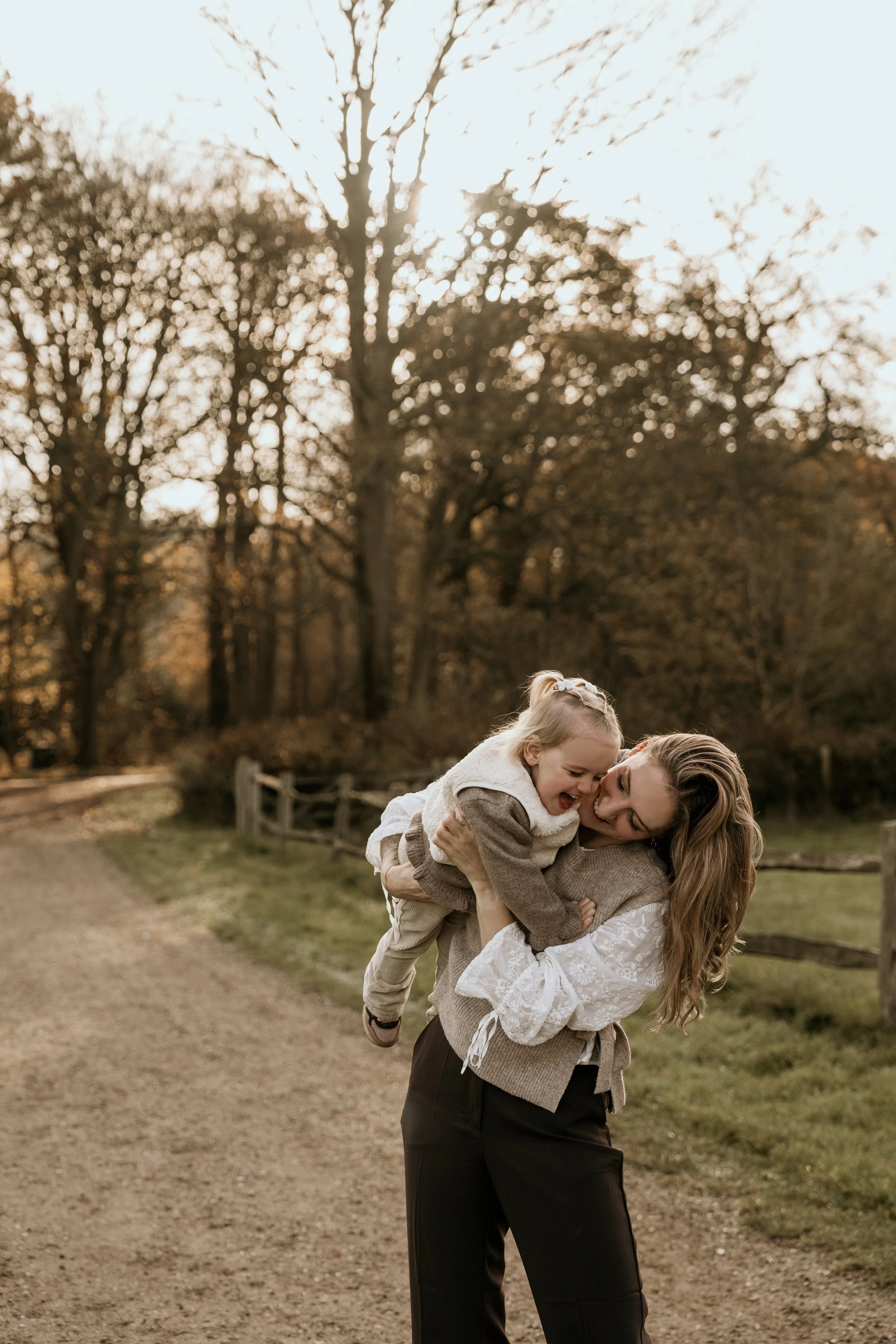 Moeder speelt met haar lachende dochter op haar schouders in een park met bomen en een pad op de achtergrond, tijdens een motherhood shoot van Rosa Lynn Signer.