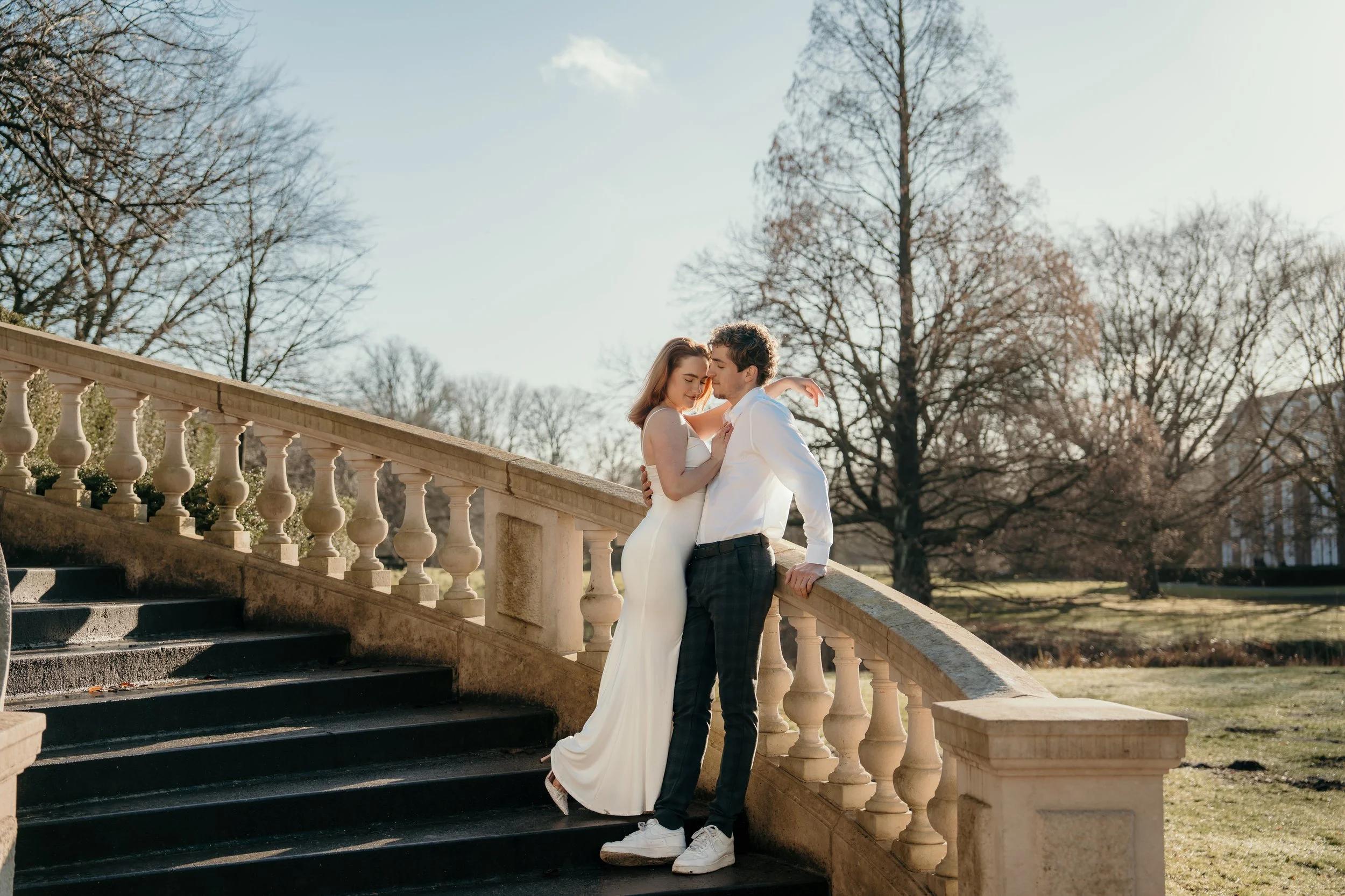 Een bruidspaar in hun trouwkleding staat dicht bij elkaar op een trap omgeven door bomen in een park, onder een heldere hemel tijdens een bruidsreportage van Rosa Lynn Signer.