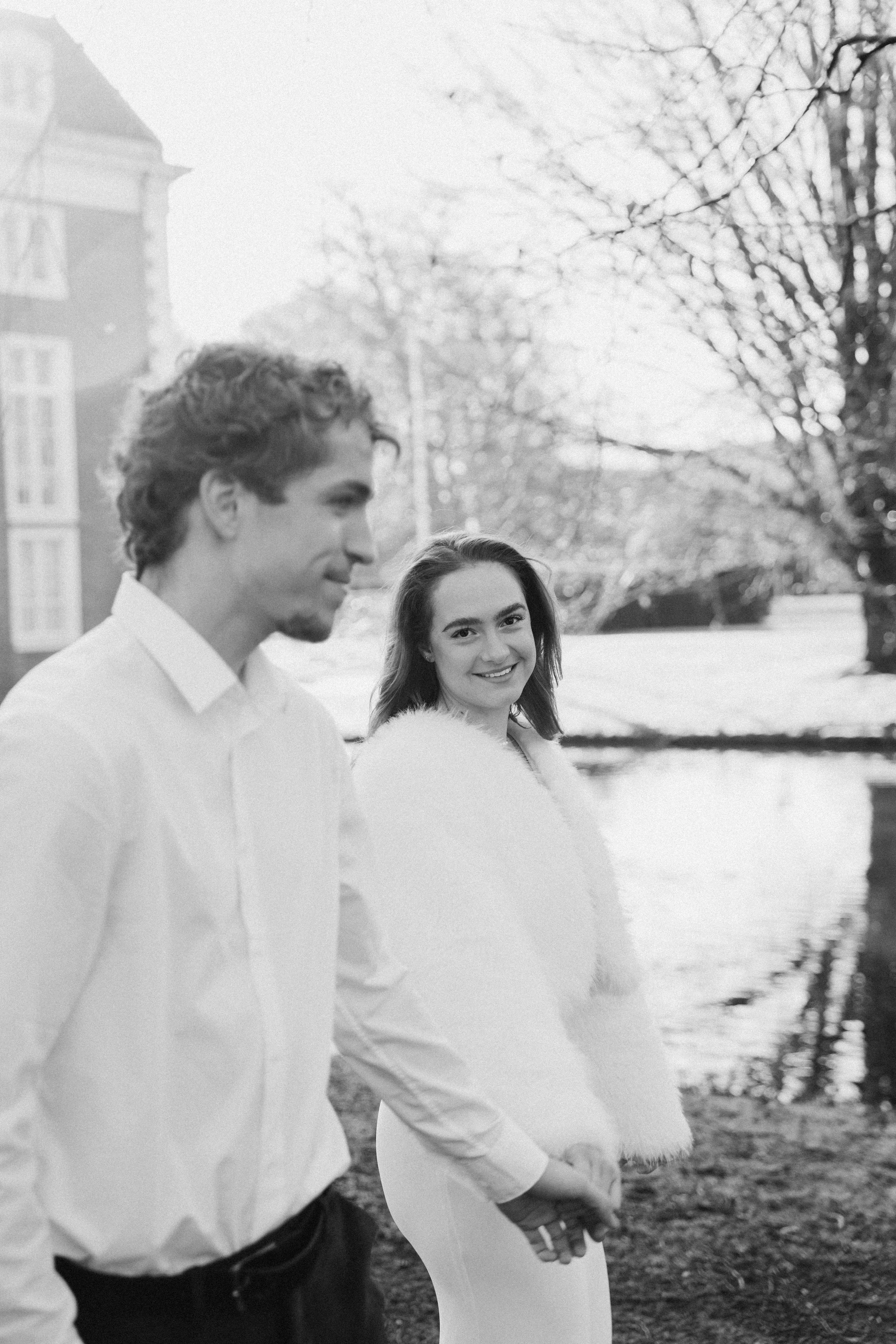 Een zwart-wit foto van een jonge bruidegom en bruid bij een meer, de vrouw glimlacht naar de camera, de man kijkt weg, in een park met bladeren aan de bomen tijdens een bruidsreportage van Rosa Lynn Signer.