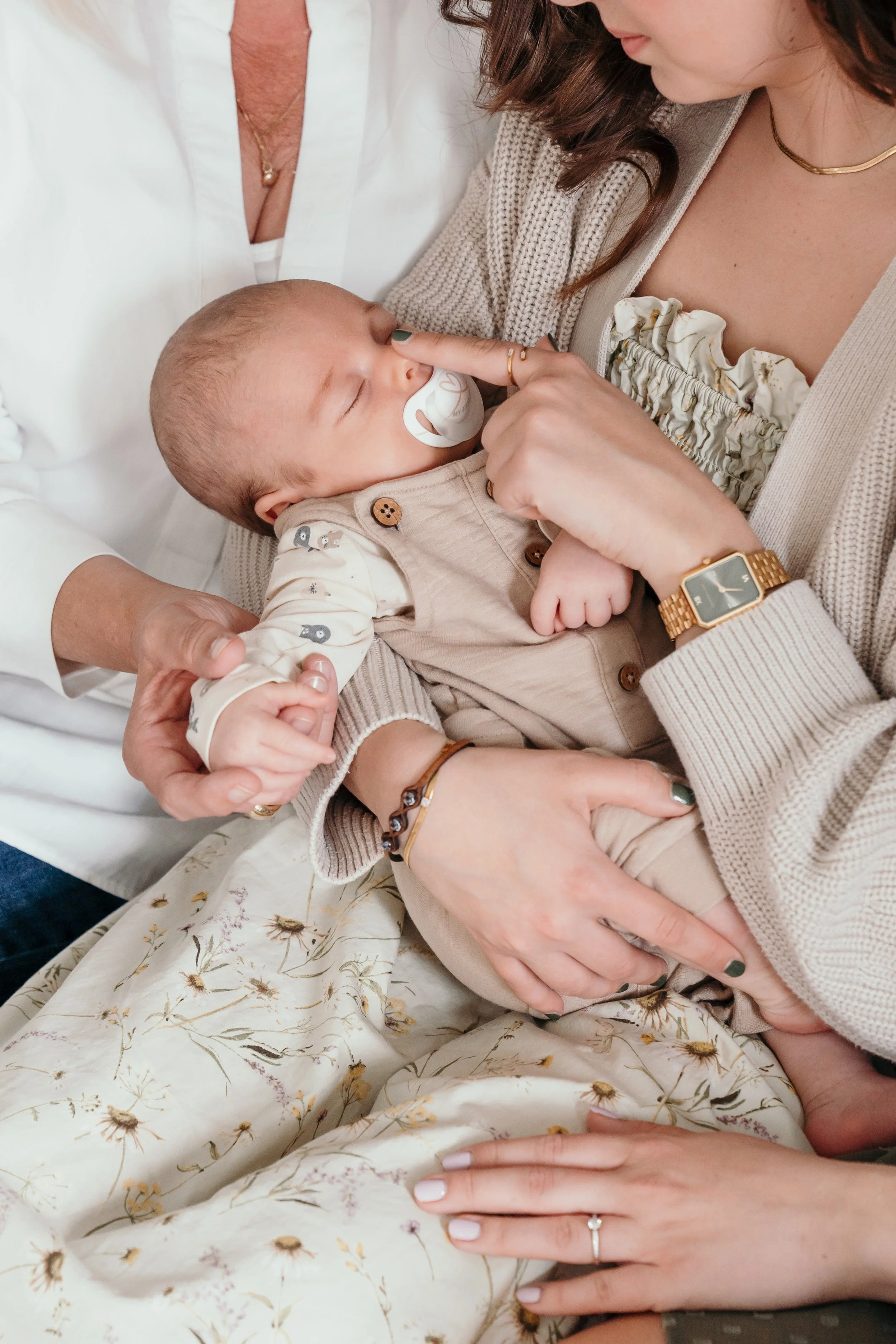 Een baby, new born, met een speentje slaapt terwijl een vrouw hem aan de neus aanraakt. Twee andere volwassenen houden de baby vast en ondersteunen hem, allemaal in een zachte, huiselijke setting tijdens een new life shoot van Rosa Lynn Signer. 