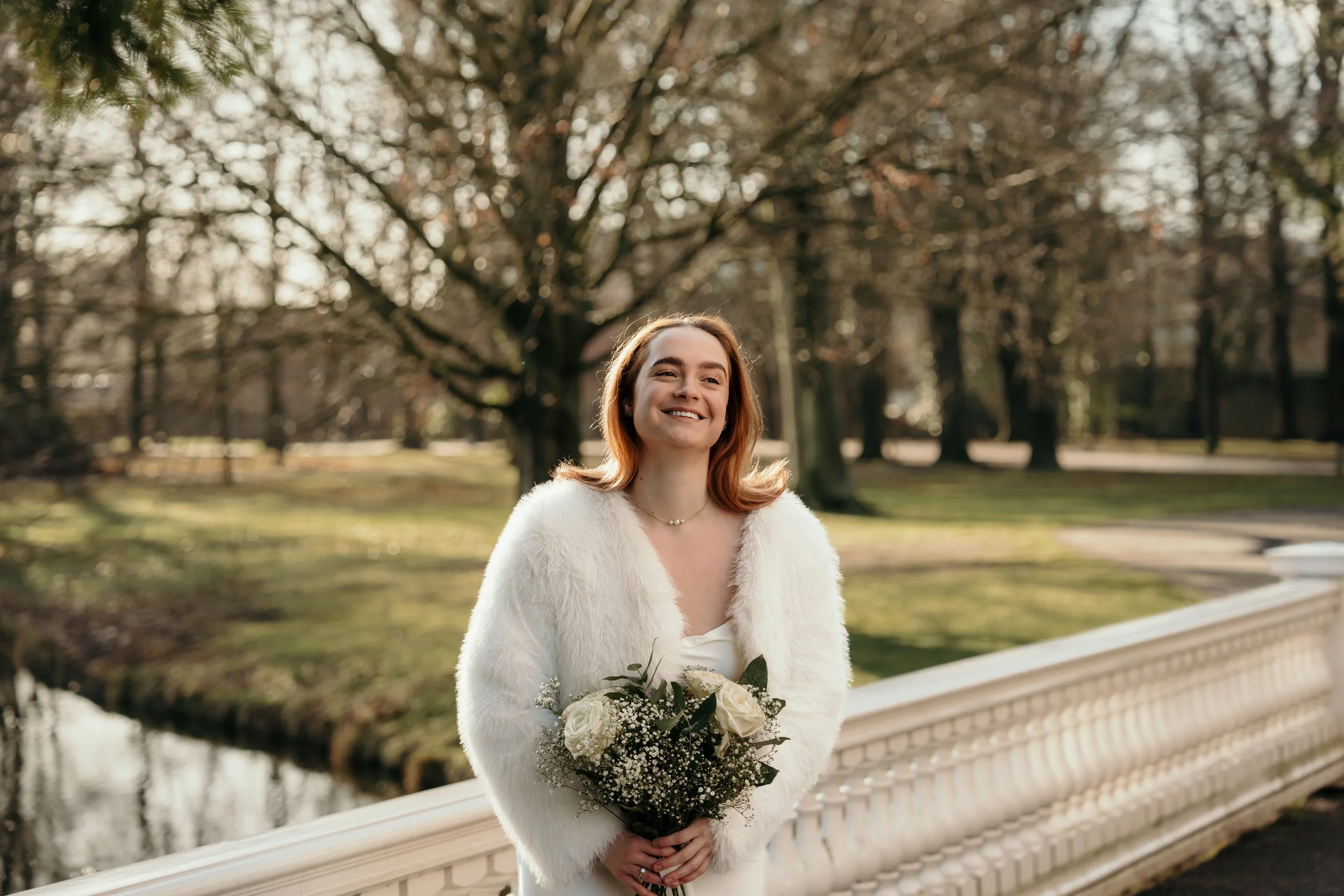 Bruid in trouwjurk en jas met bruidsboekt tussen een park met bomen en een kleine rivier tijdens een bruidsreportage van Rosa Lynn Signer.