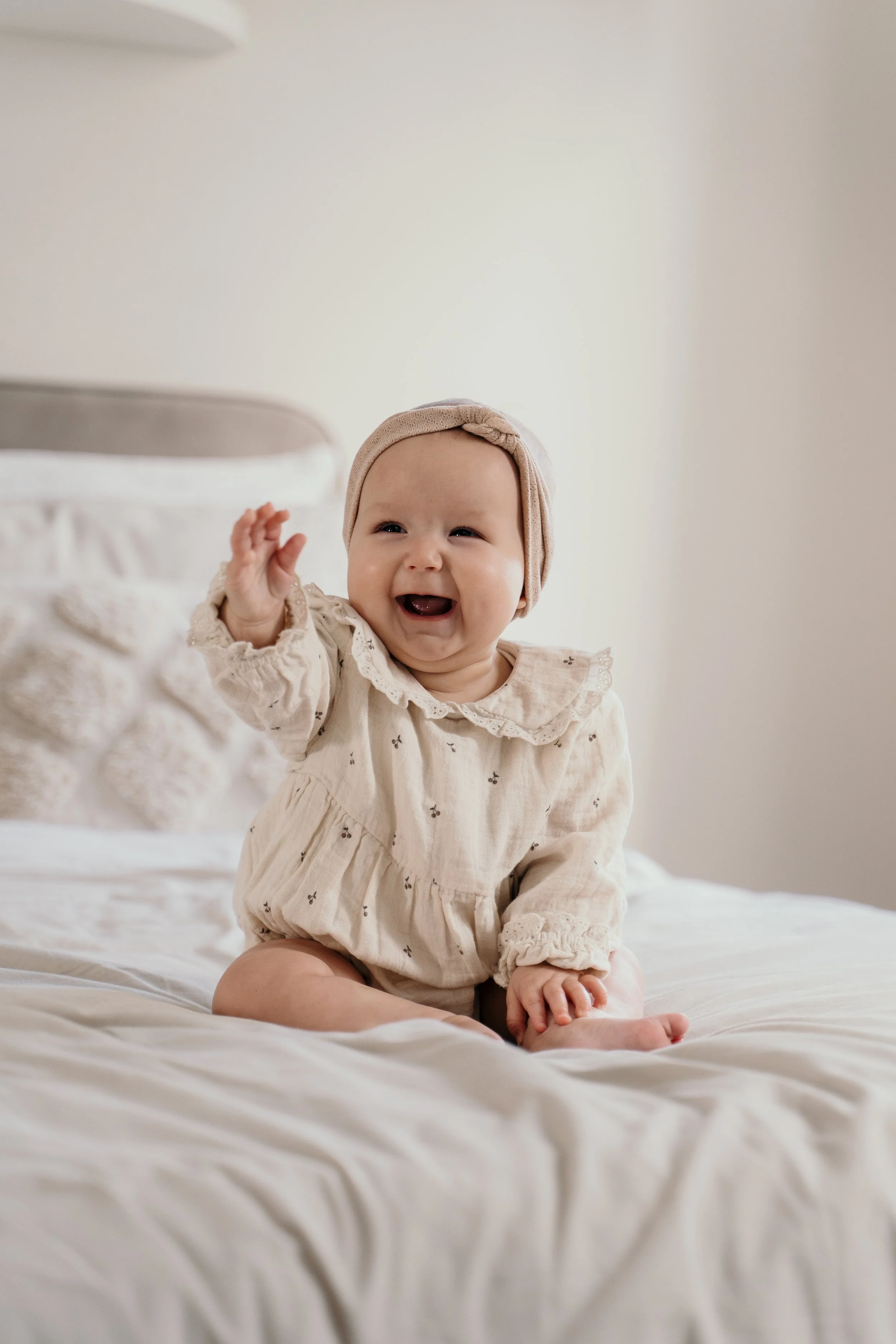 Lachende baby in beige outfit en hoofdbandje, zittend op een wit bed, vrolijk blij tijdens een new life shoot van Rosa Lynn Signer. 