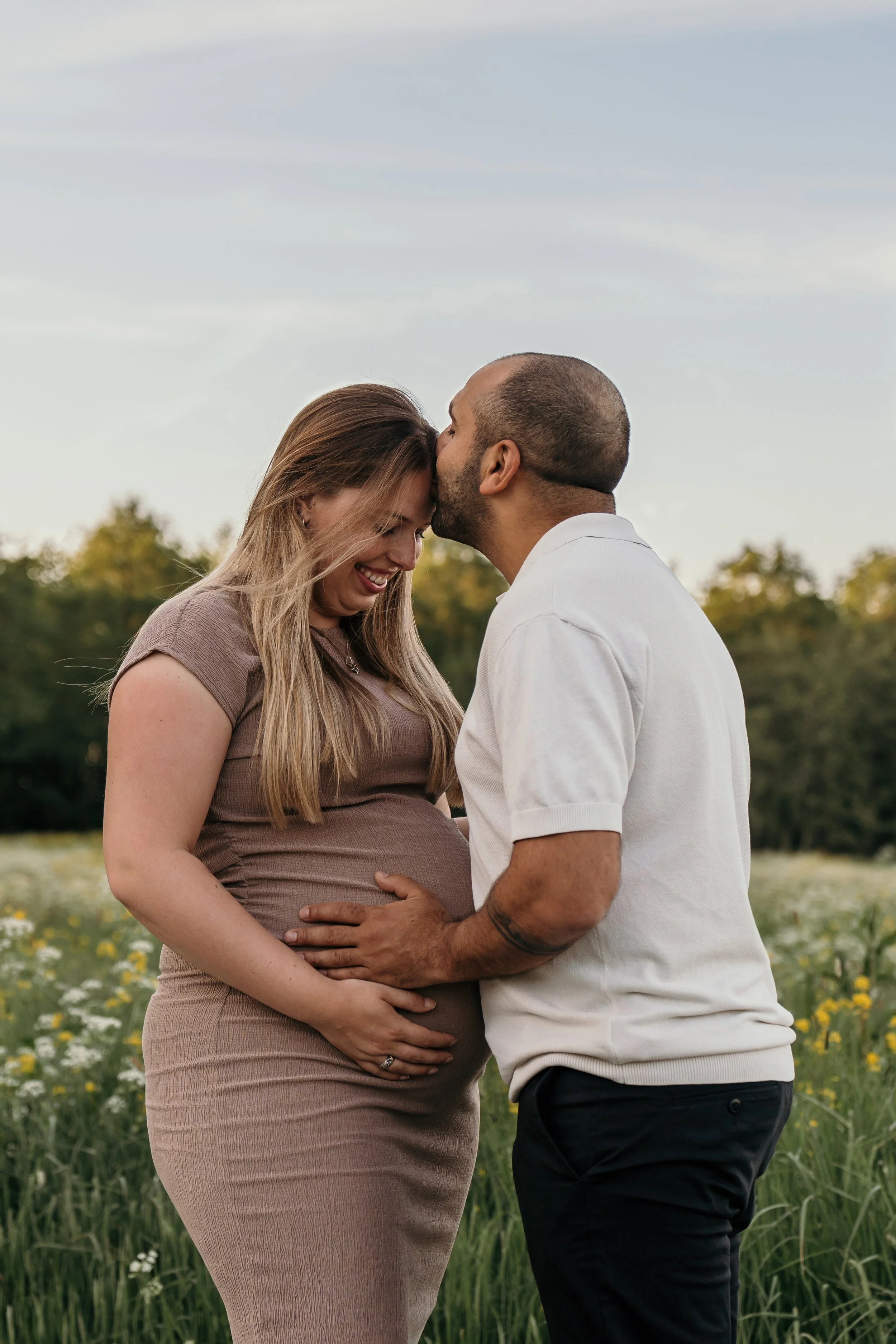 Een zwangere vrouw en haar partner staan in een veld met bloemen, ze delen een liefdevolle knuffel en glimlachen naar elkaar tijdens een zwangerschapsshoot van Rosa Lynn Signer.