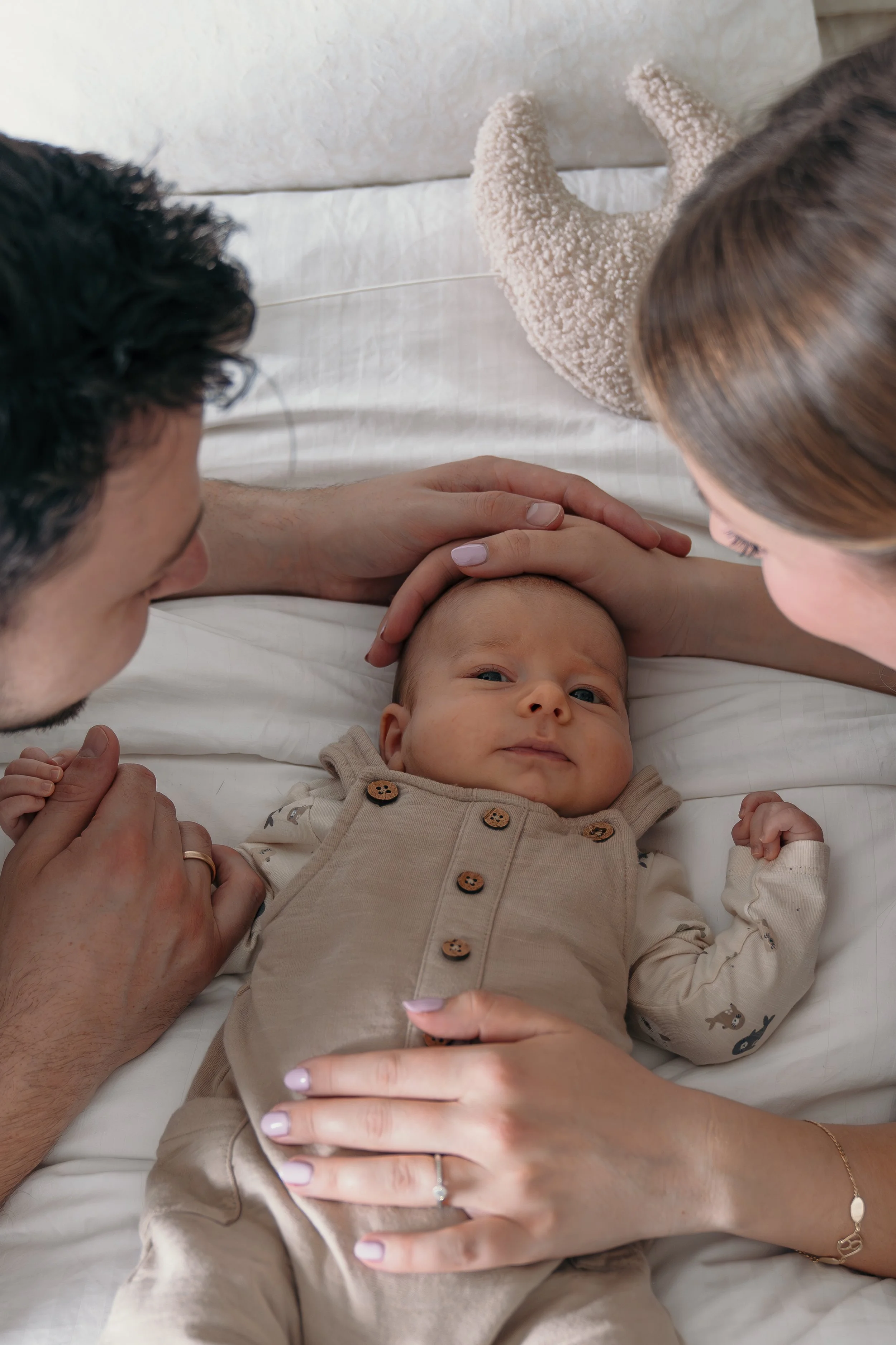 Een baby ligt op een bed, omringd door zijn mama en papa die naar hem kijken en hem liefdevol aanraken tijdens een new born shoot van Rosa Lynn Signer.