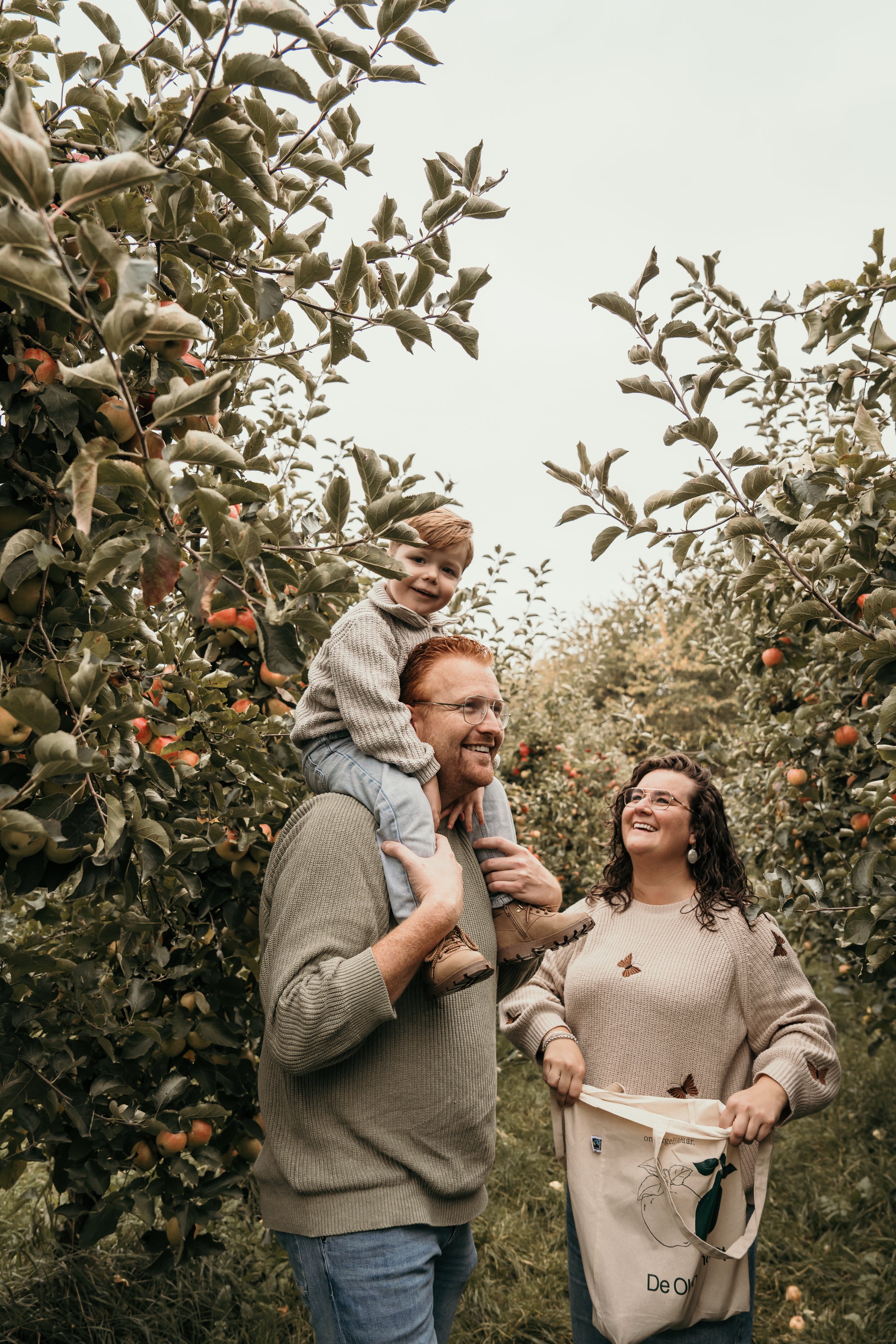 Gezin in een appelboomgaard, vader draagt een jongen op zijn schouders, moeder staat naast hen met een boodschappentas, lachen en genieten van een dag in de natuur tijdens een gezinsshoot van Rosa Lynn Signer.