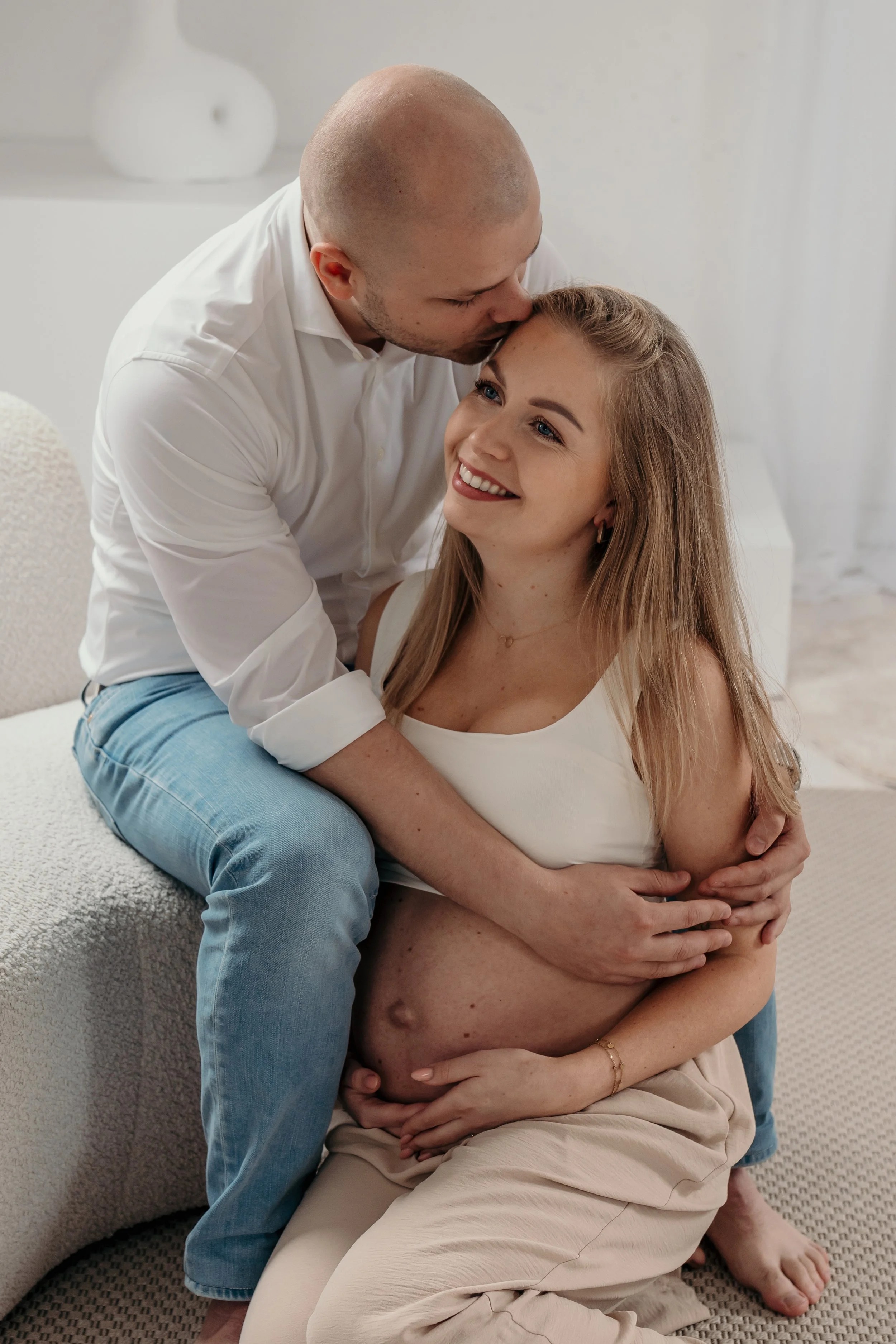 Een gelukkige zwangere vrouw zit op haar knieën en houdt haar buik, terwijl een man haar van achteren omhelst en haar kust op het voorhoofd tijdens een zwangerschapsshoot van Rosa Lynn Signer in een daglichstudio.