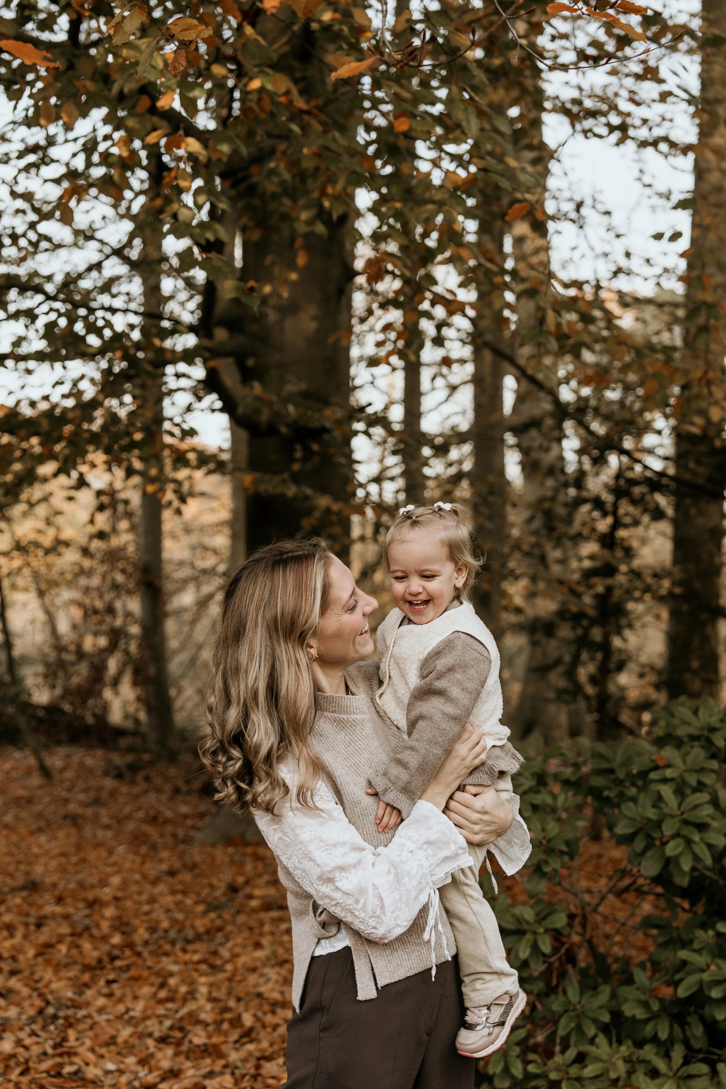 Moeder houdt haar lachende dochter in haar armen in een bos met herfstkleuren tijdens een motherhood shoot van Rosa Lynn Signer.