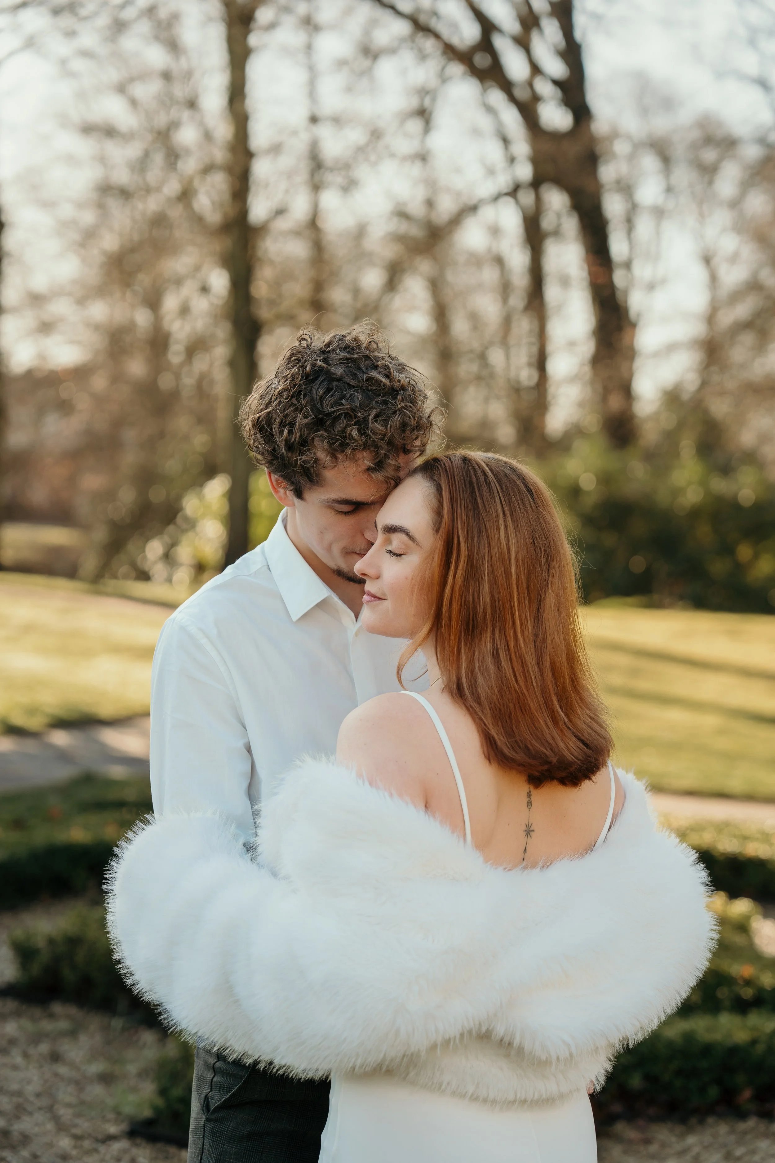 Een bruid en bruidegom knuffelen elkaar liefdevol in een park, de man heeft krullig haar en de vrouw heeft rood haar en draagt een witte bruidsjurk en bontjas tijdens een bruidsreportage van Rosa Lynn Signer.