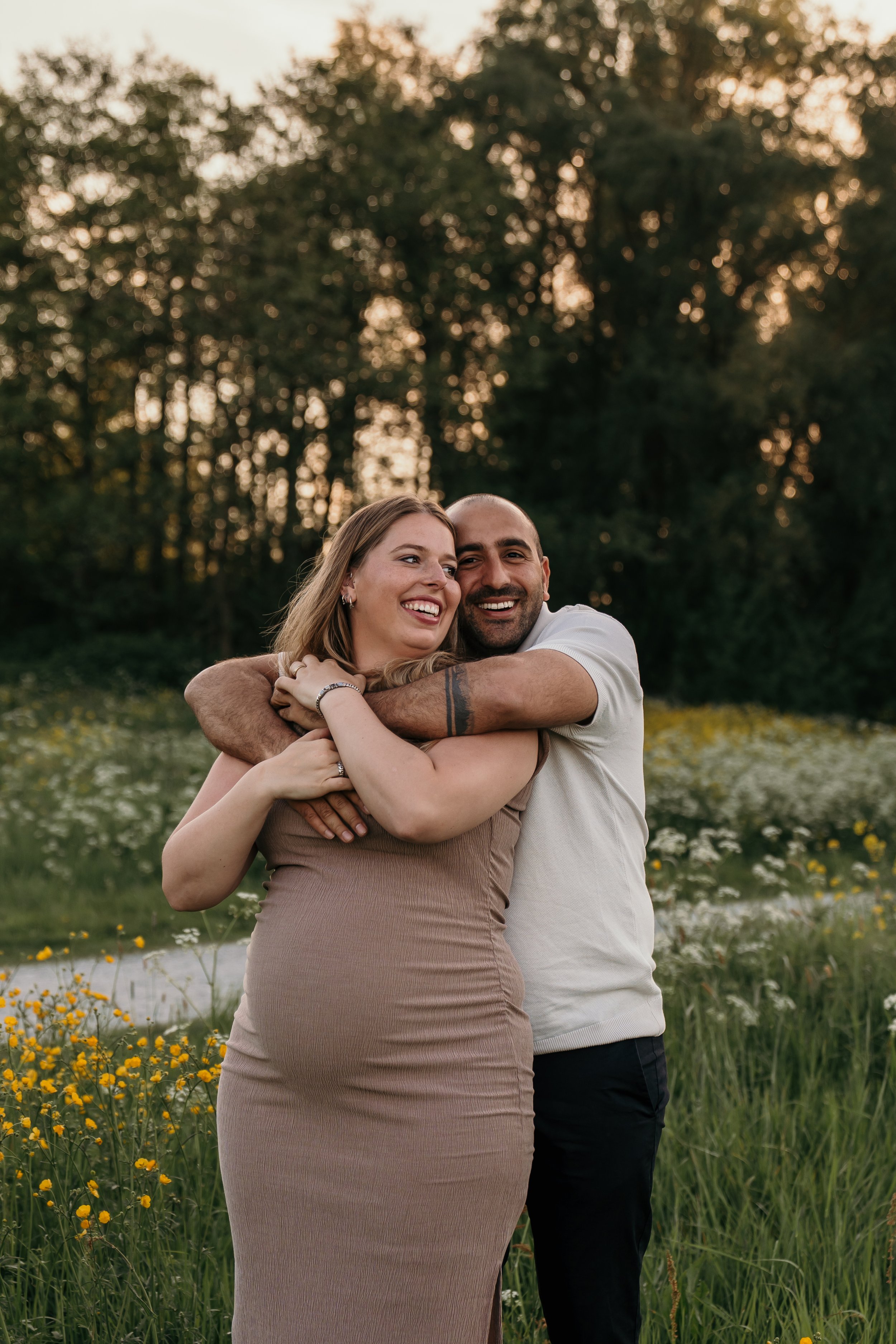 Een lachend paar dat elkaar omhelst in een veld met bloemen tijdens zonsondergang tijdens een zwangerschapsshoot van Rosa Lynn Signer.