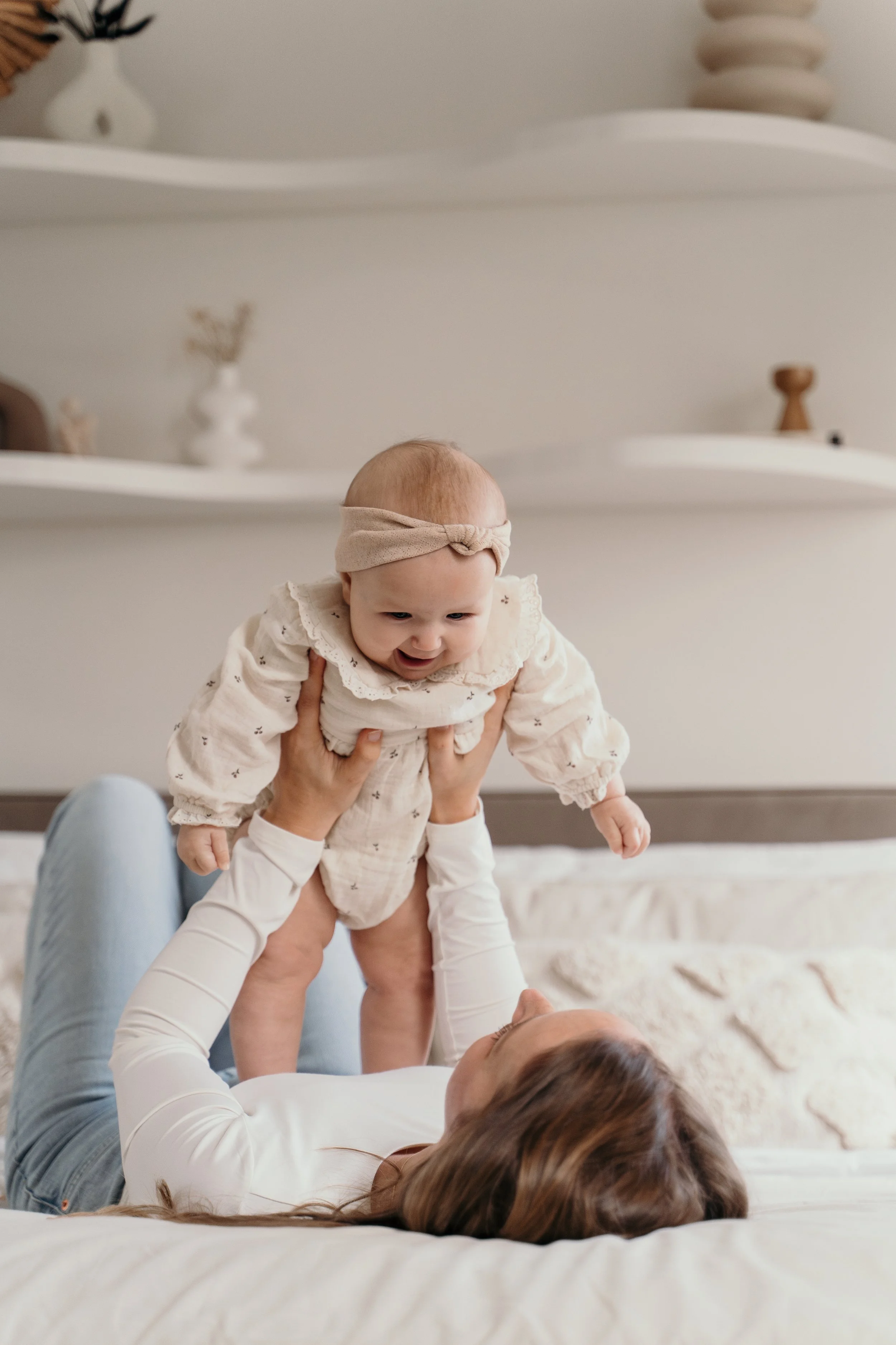 Een baby die door haar moeder omhoog wordt gehouden terwijl ze op een bed ligt in een slaapkamer met witte planken en decoratieve voorwerpen op de achtergrond tijdens een motherhood / new born shoot van Rosa Lynn Signer in een studio.