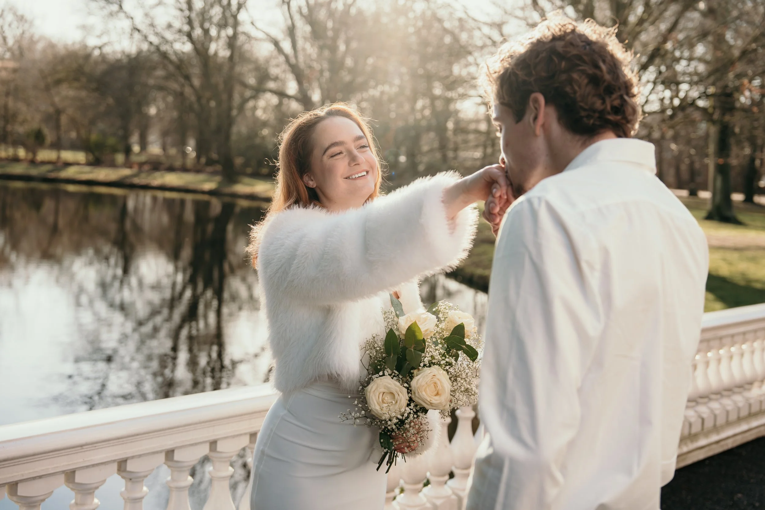 Een bruid met in haar bruidsjurk en de bruidegom kust haar trouwring bij een meer met een brug en bomen op de achtergrond tijdens een bruidsreportage van Rosa Lynn Signer..