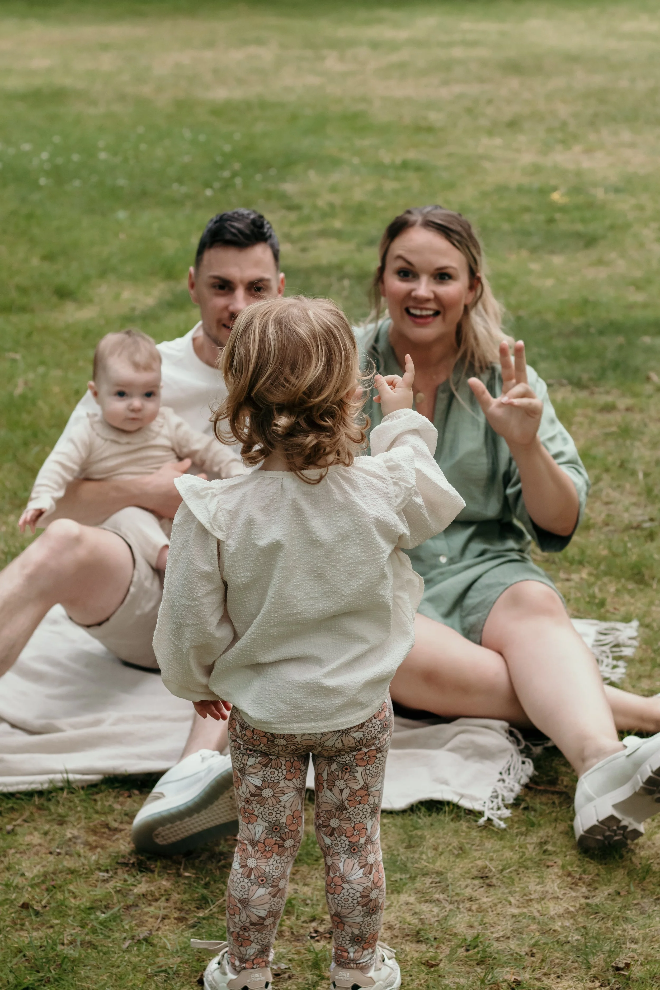 Een gezin speelt op het gras in een park; ouders zitten op een deken met hun twee jonge kinderen, een meisje staat en interacteert met de ouders tijdens een gezinsshoot van Rosa Lynn Signer.