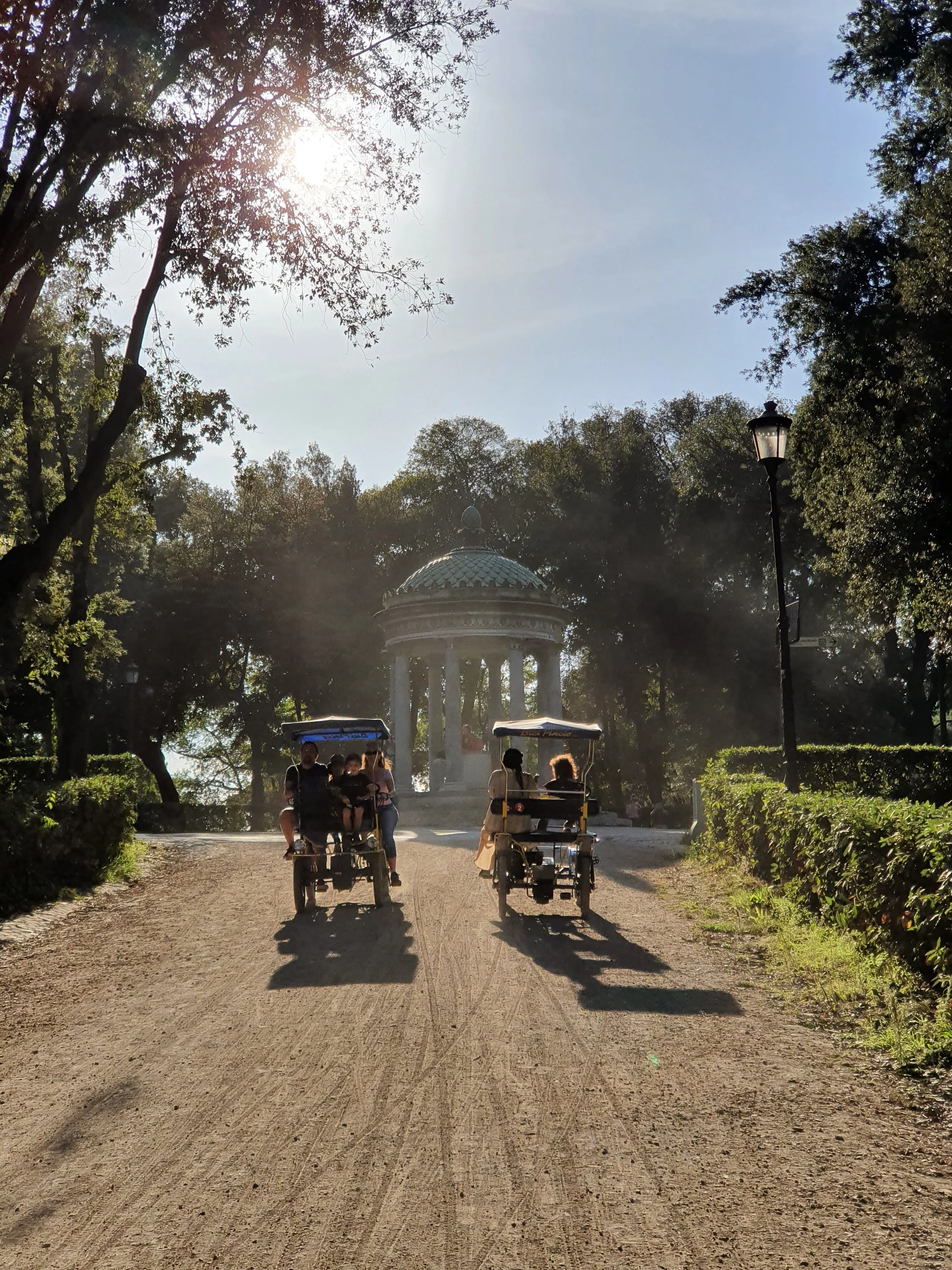 Twee golfkarretjes met mensen rijden op een zandweg in een park met bomen en een kenotafon in de achtergrond. Tijdens een zonnige dag.