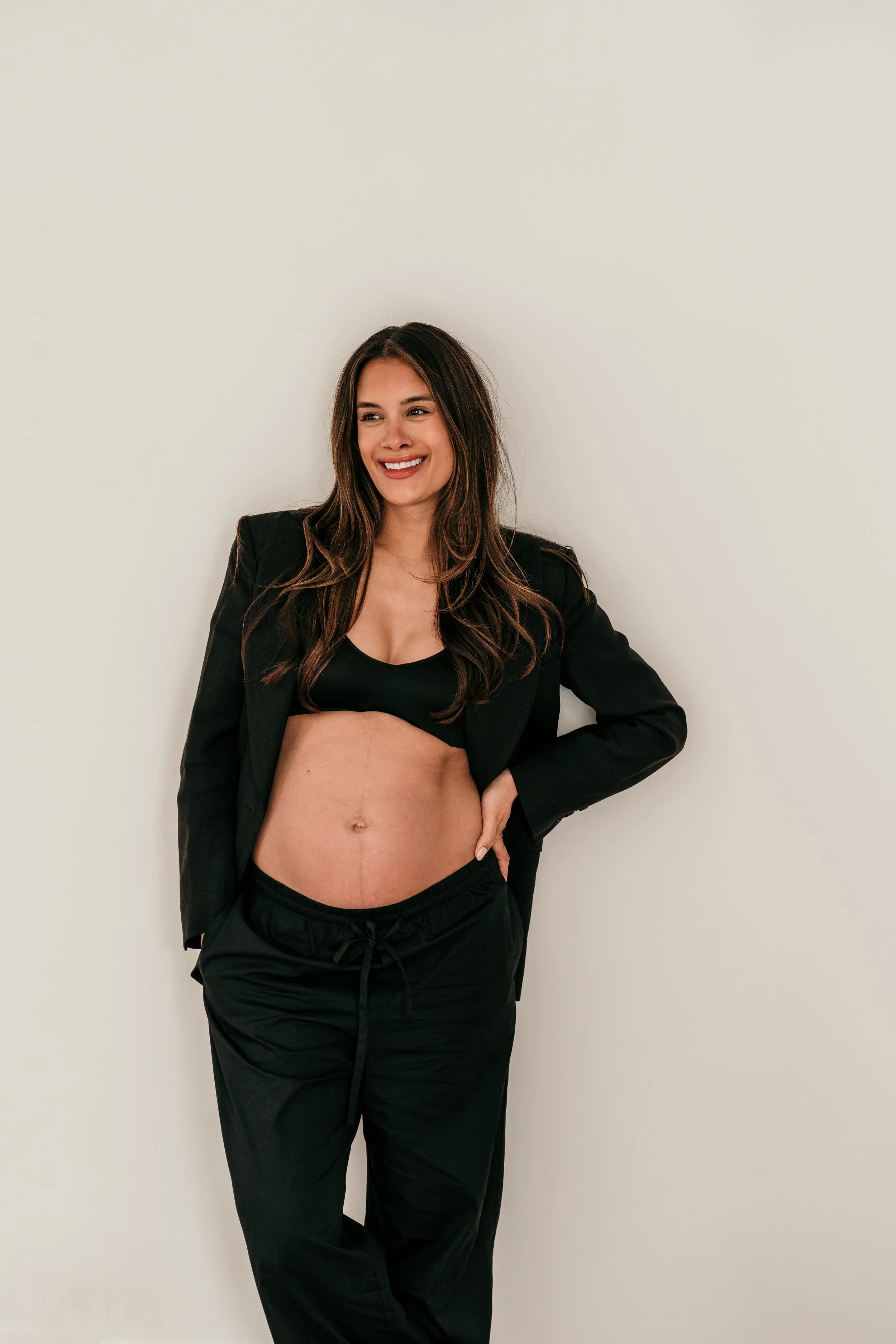 Zwangere vrouw leunt tegen een witte muur en glimlacht tijdens een zwangerschapsshoot van Rosa Lynn Signer.