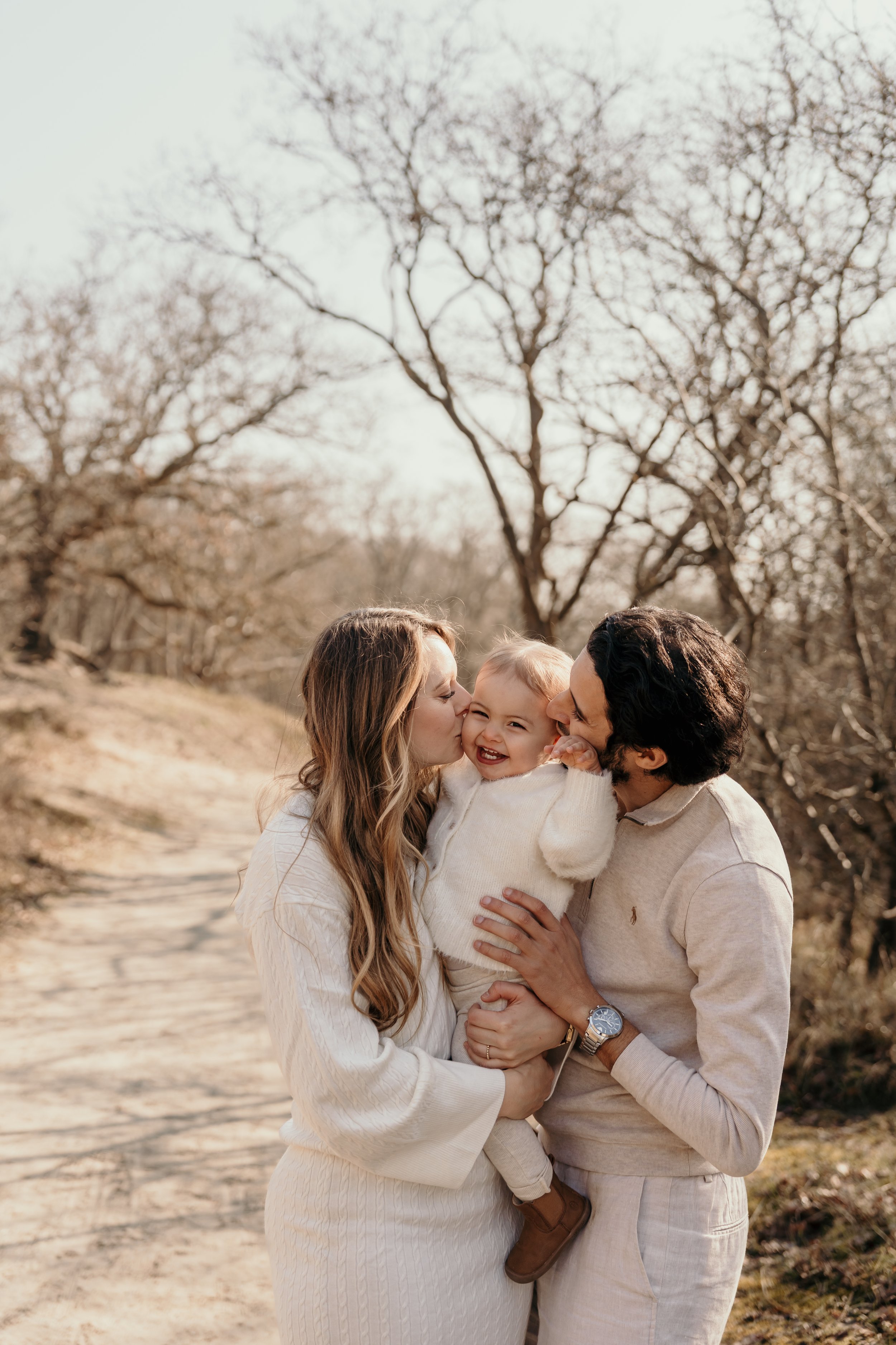 Gezinsshoot in de duinen. Kindje lacht terwijl de ouders een kusje aan haar geven tijdens een fotoshoot van Rosa Lynn Signer
