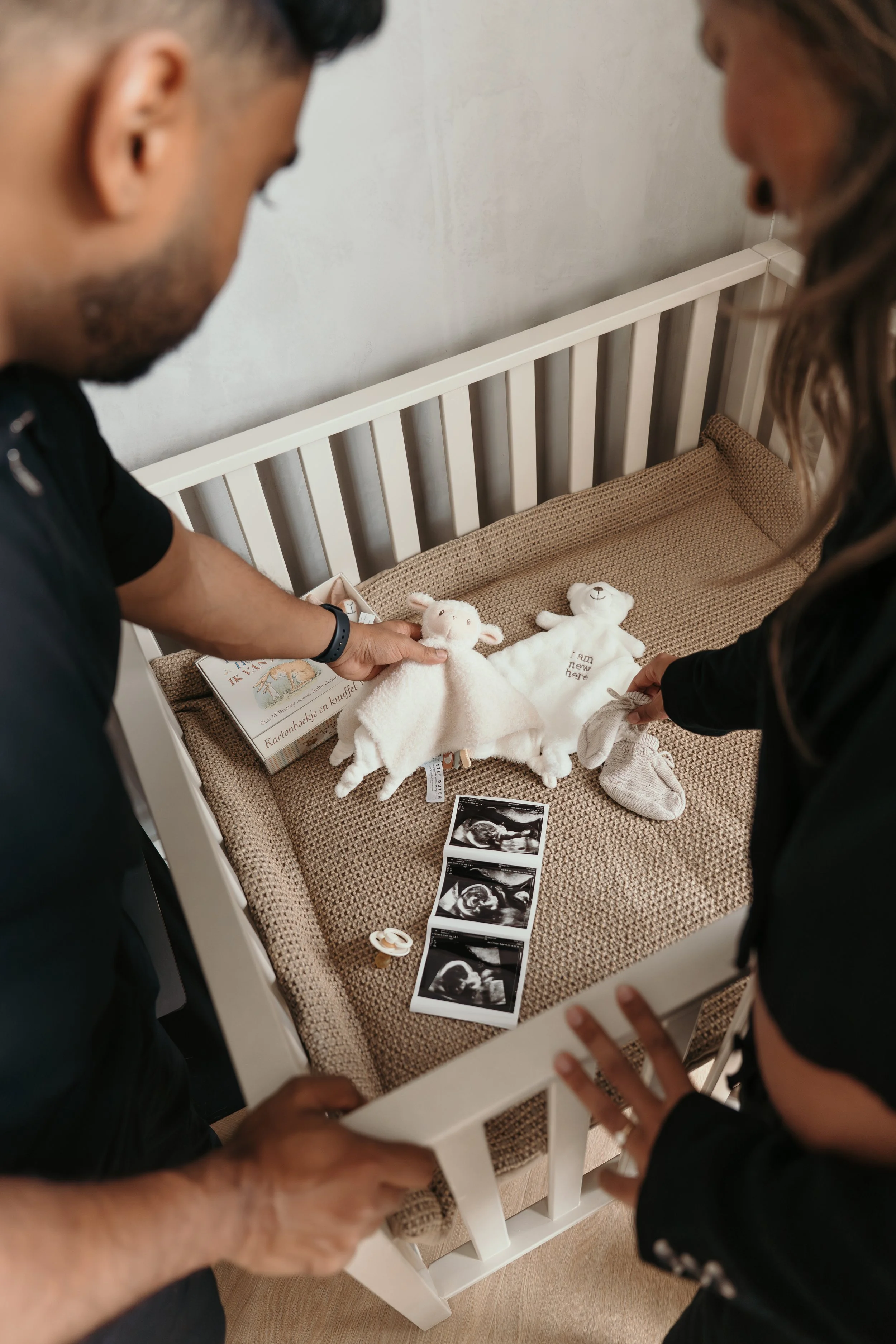 Twee nieuwe ouders bekijken babyspullen en echofoto's in een wiegje met geboortekleding, knuffels en gezonde babyfoto's tijdens een zwangerschapsshoot van Rosa Lynn Signer.